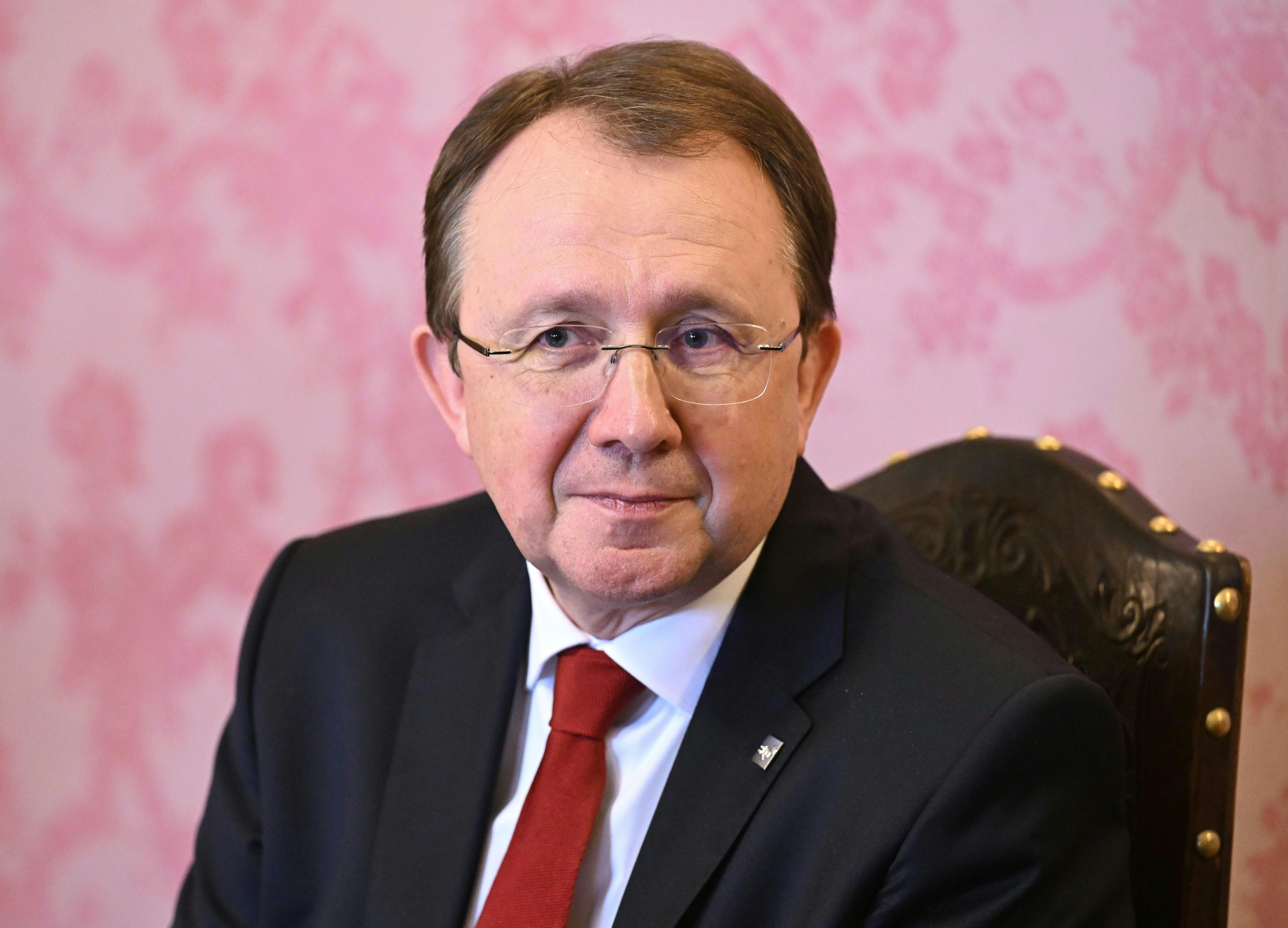 Ein Mann mit Brille im Anzug und roter Krawatte sitzt vor rosa gemustertem Hintergrund.