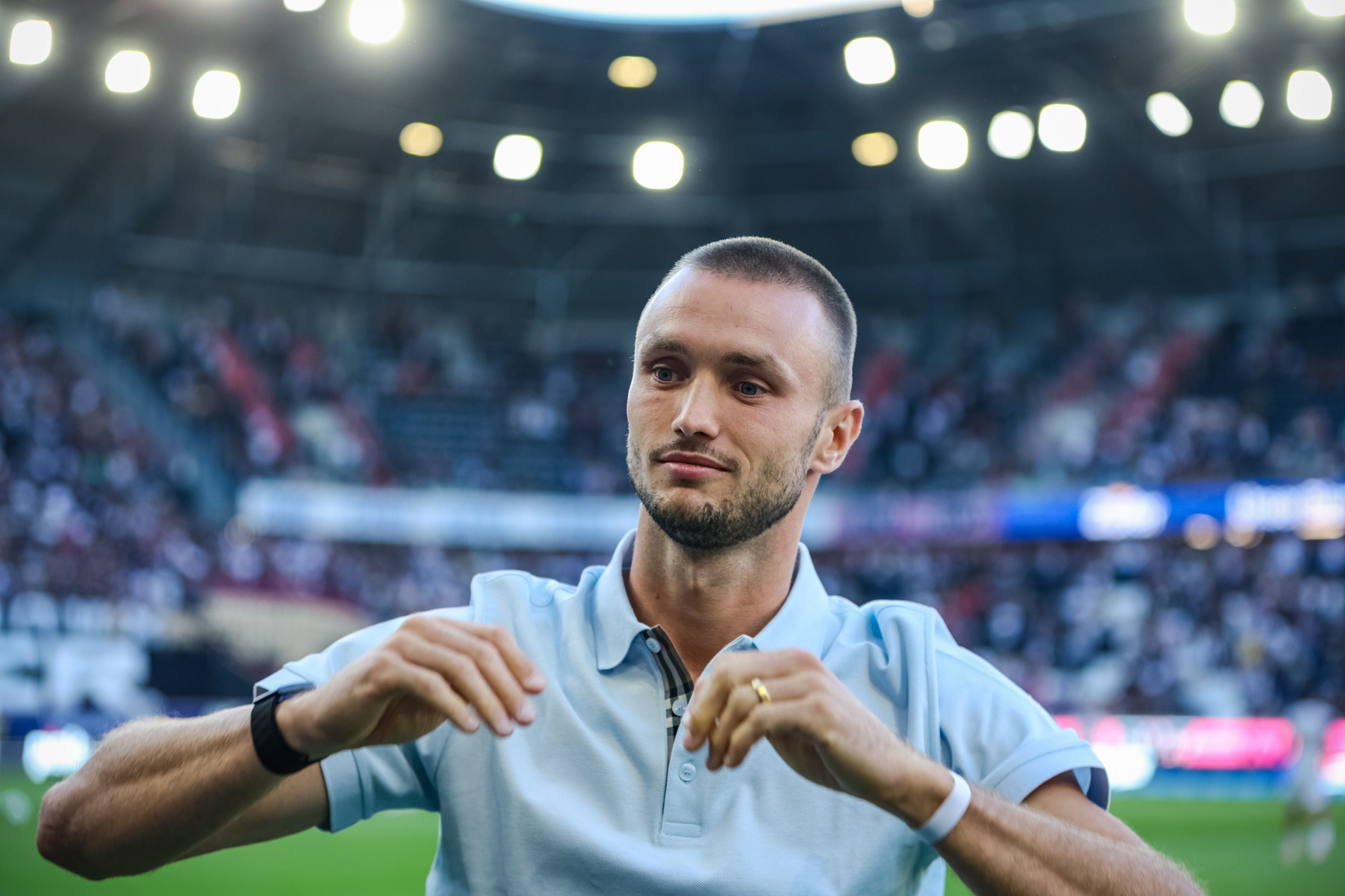 Mann im hellblauen Polohemd steht im Fußballstadion vor unscharfem Publikum.