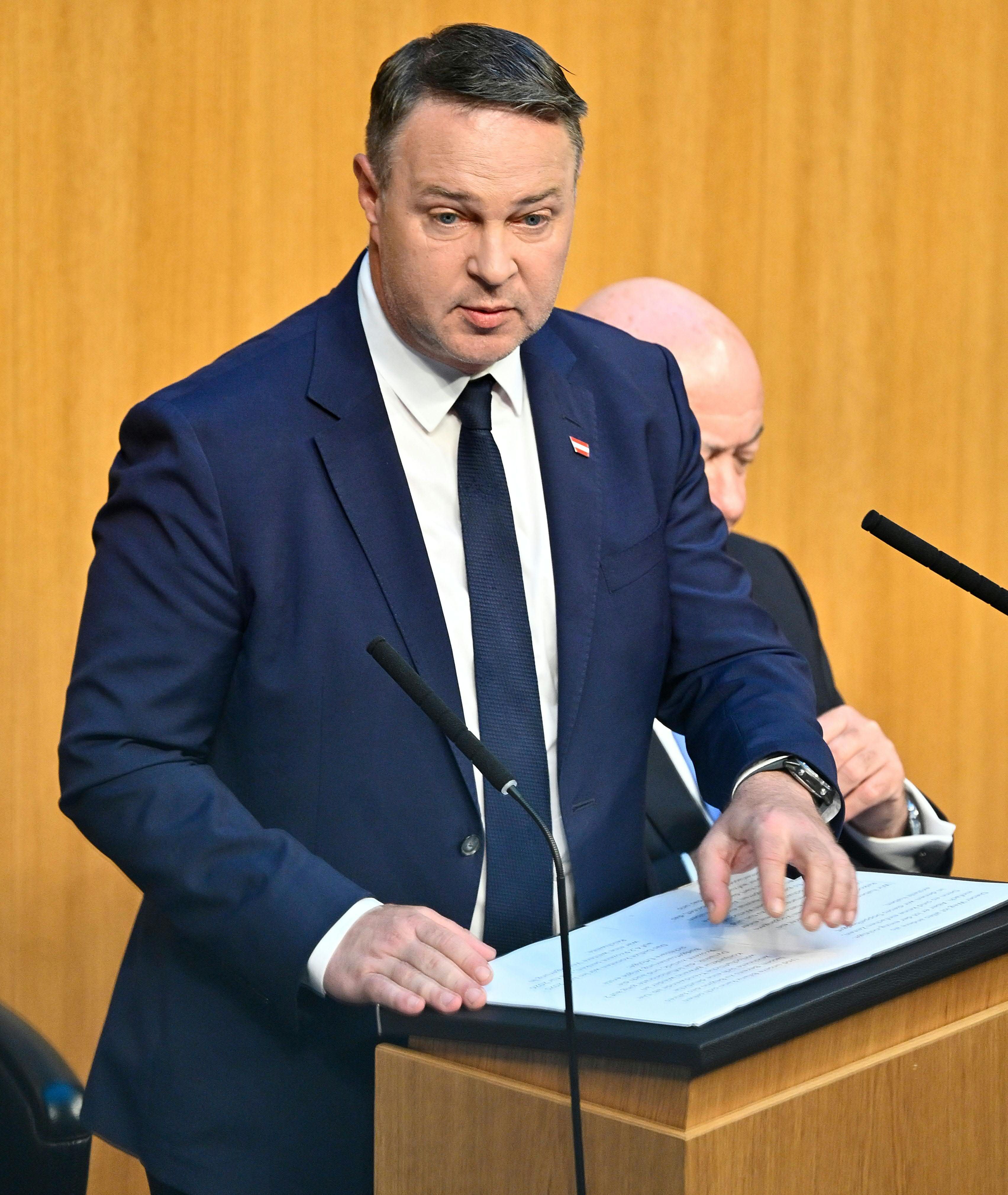 Ein Mann im Anzug spricht am Rednerpult im Parlament vor Mikrofonen.