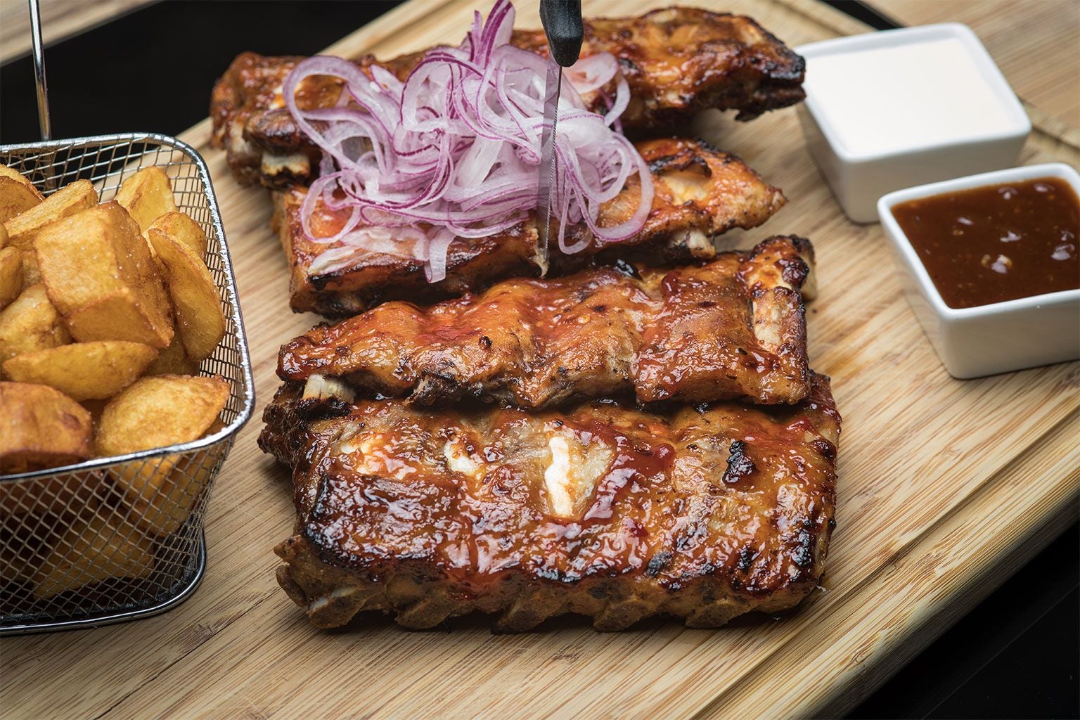 Gegrillte Spareribs mit Zwiebeln, Bratkartoffeln und zwei Dips auf einem Holzbrett serviert.