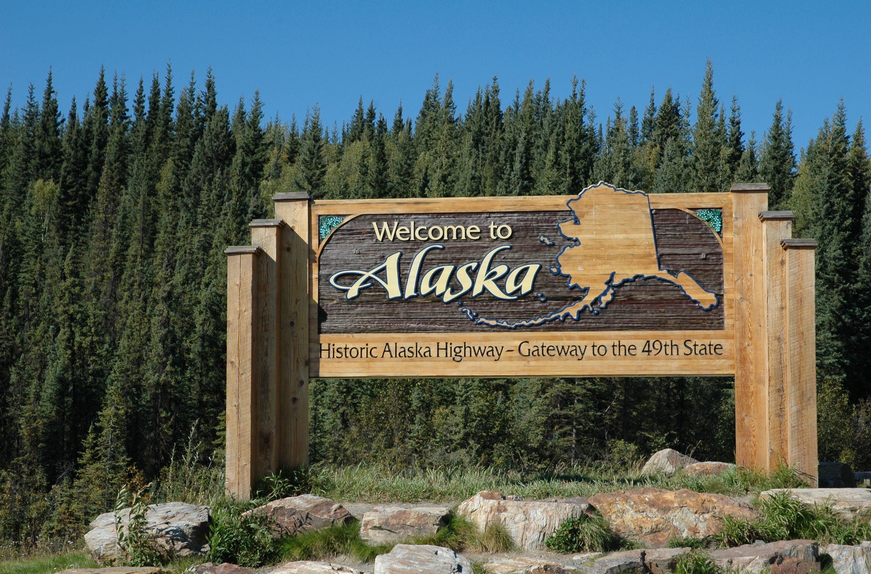 Holzschild „Welcome to Alaska“ an der Grenze mit Wald im Hintergrund.