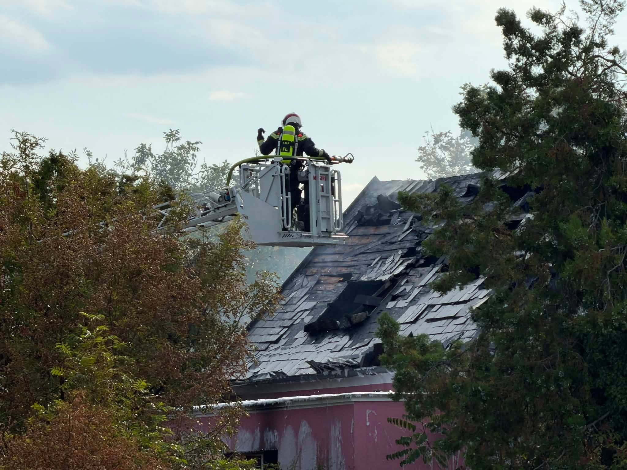 Feuerwehrmann löscht Brand auf beschädigtem Dach mit Schlauch aus einer Drehleiter.