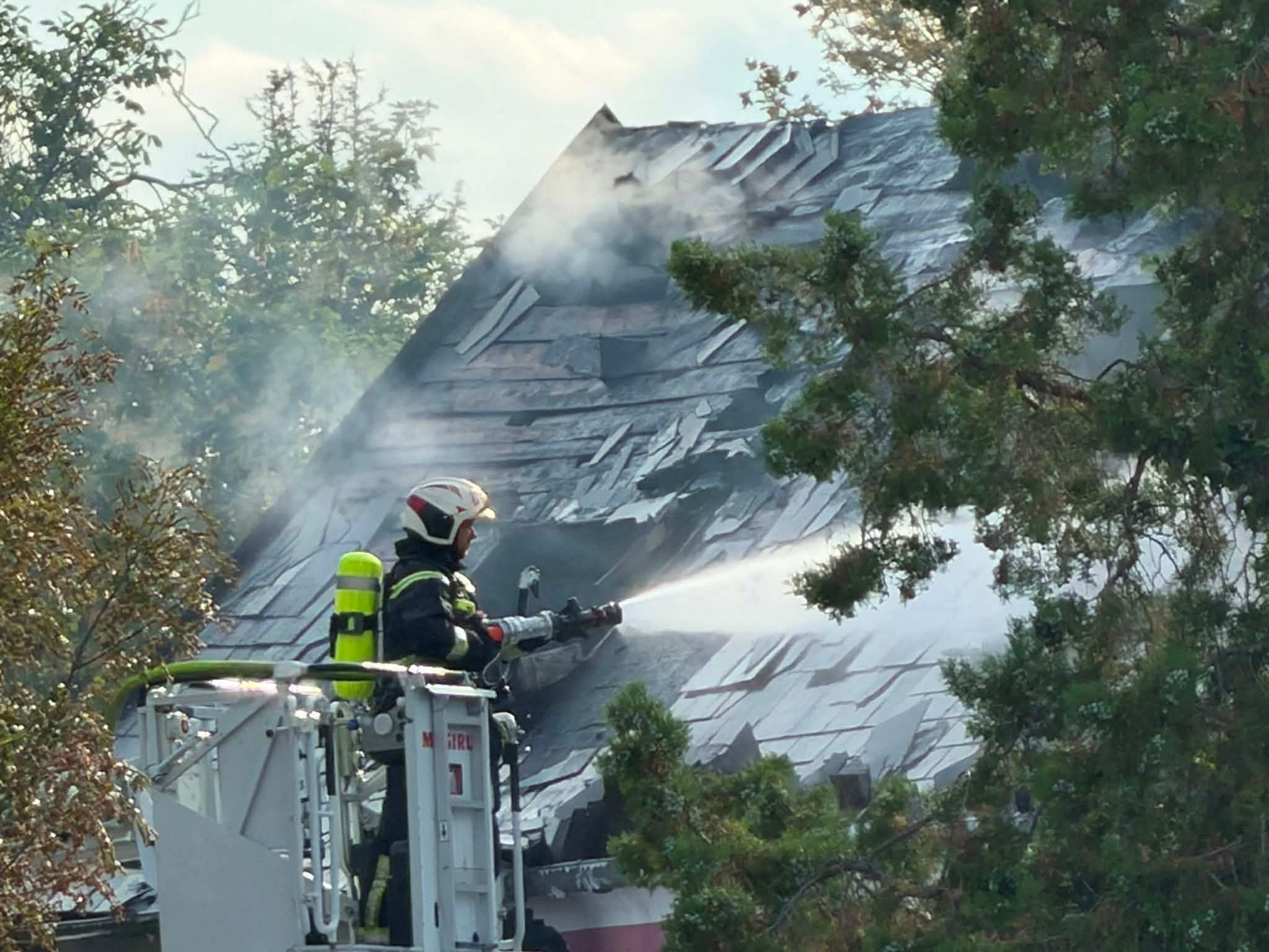 Feuerwehrmann löscht brennendes Hausdach mit Wasserstrahl, Rauch steigt auf.