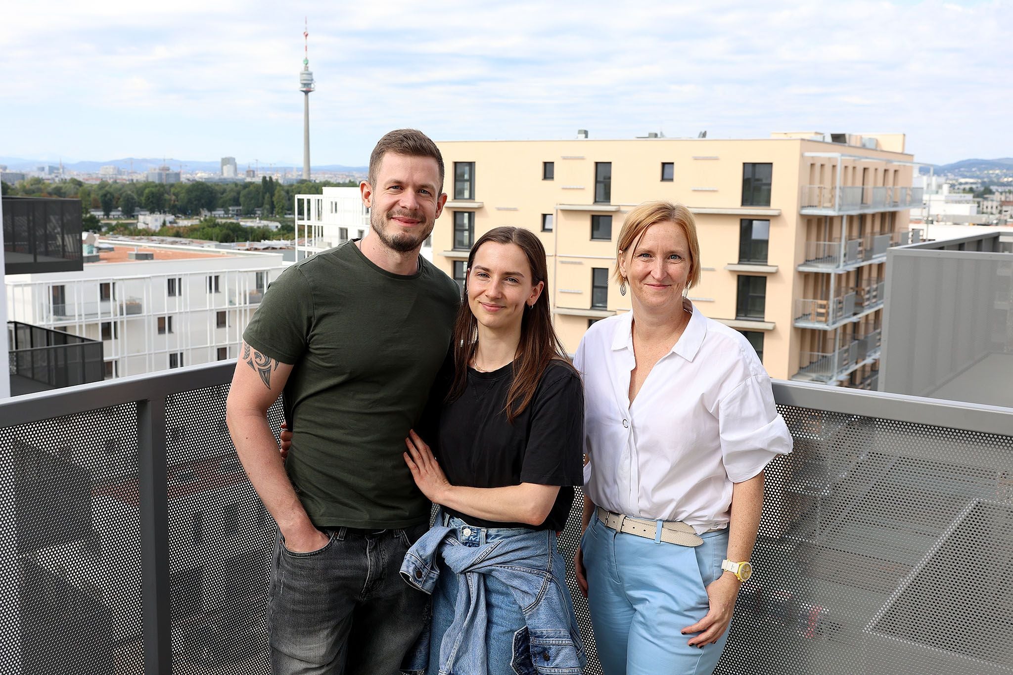 Drei Personen stehen auf einem Balkon vor modernen Wohnhäusern und lächeln in die Kamera.