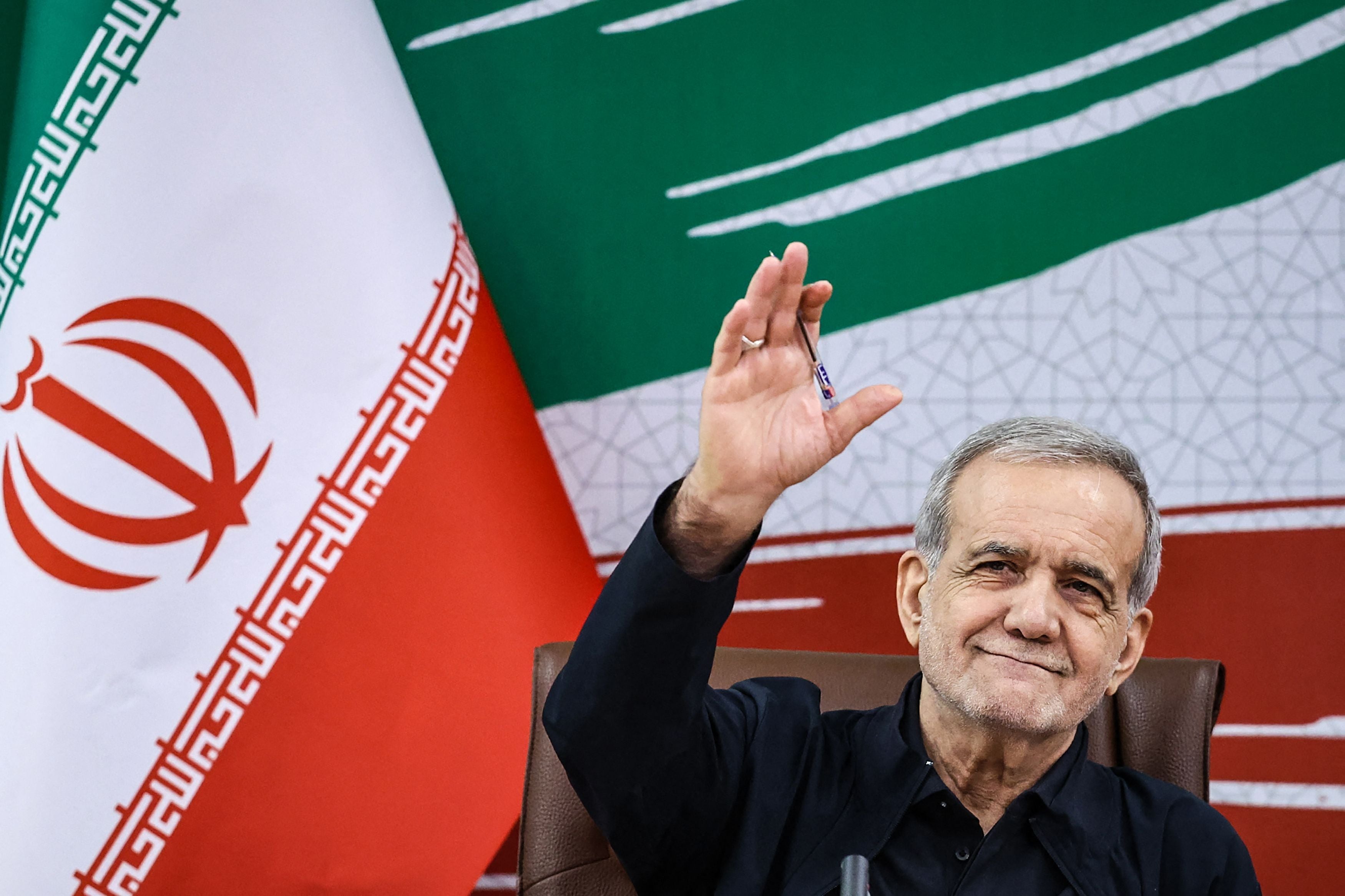 Ein Mann winkt lächelnd vor einer iranischen Flagge bei einer Pressekonferenz.