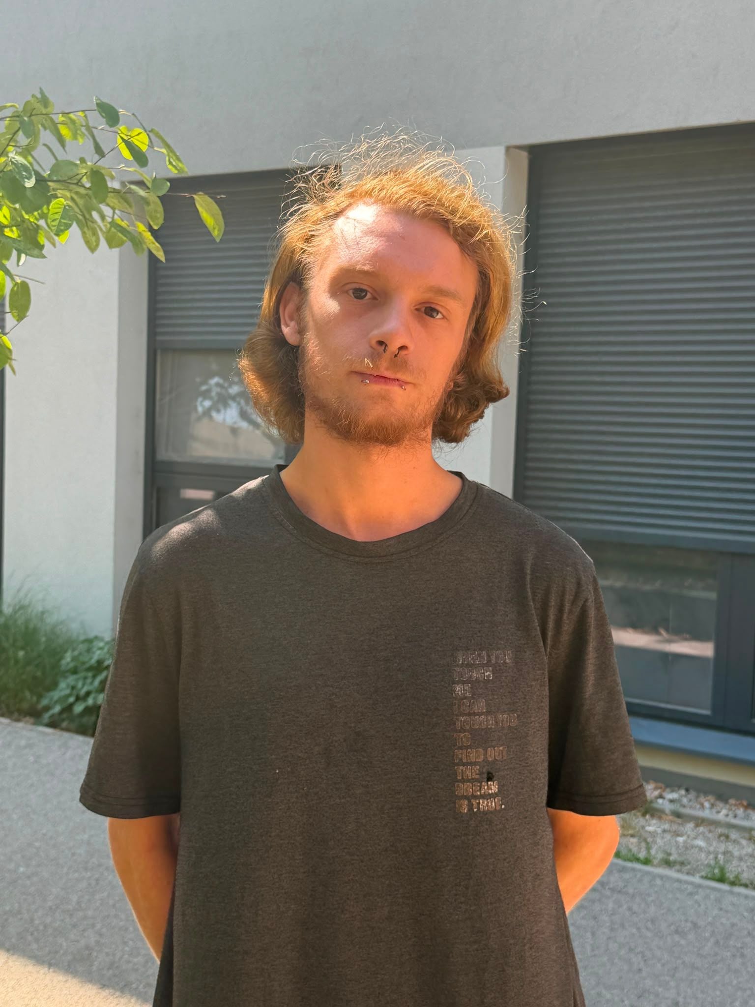 Junger Mann mit dunklem T-Shirt steht draußen vor einem Gebäude bei Sonnenschein.