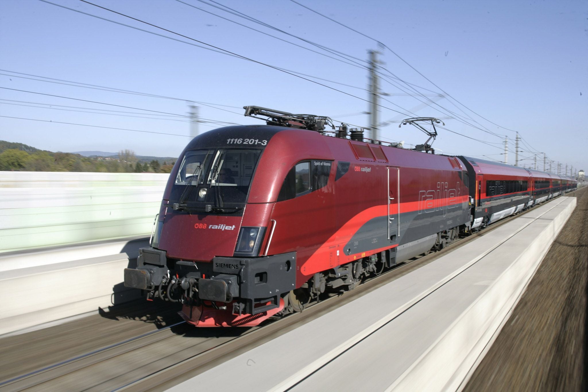 Ein roter ÖBB railjet-Schnellzug fährt auf einer modernen Bahnstrecke bei Tageslicht.