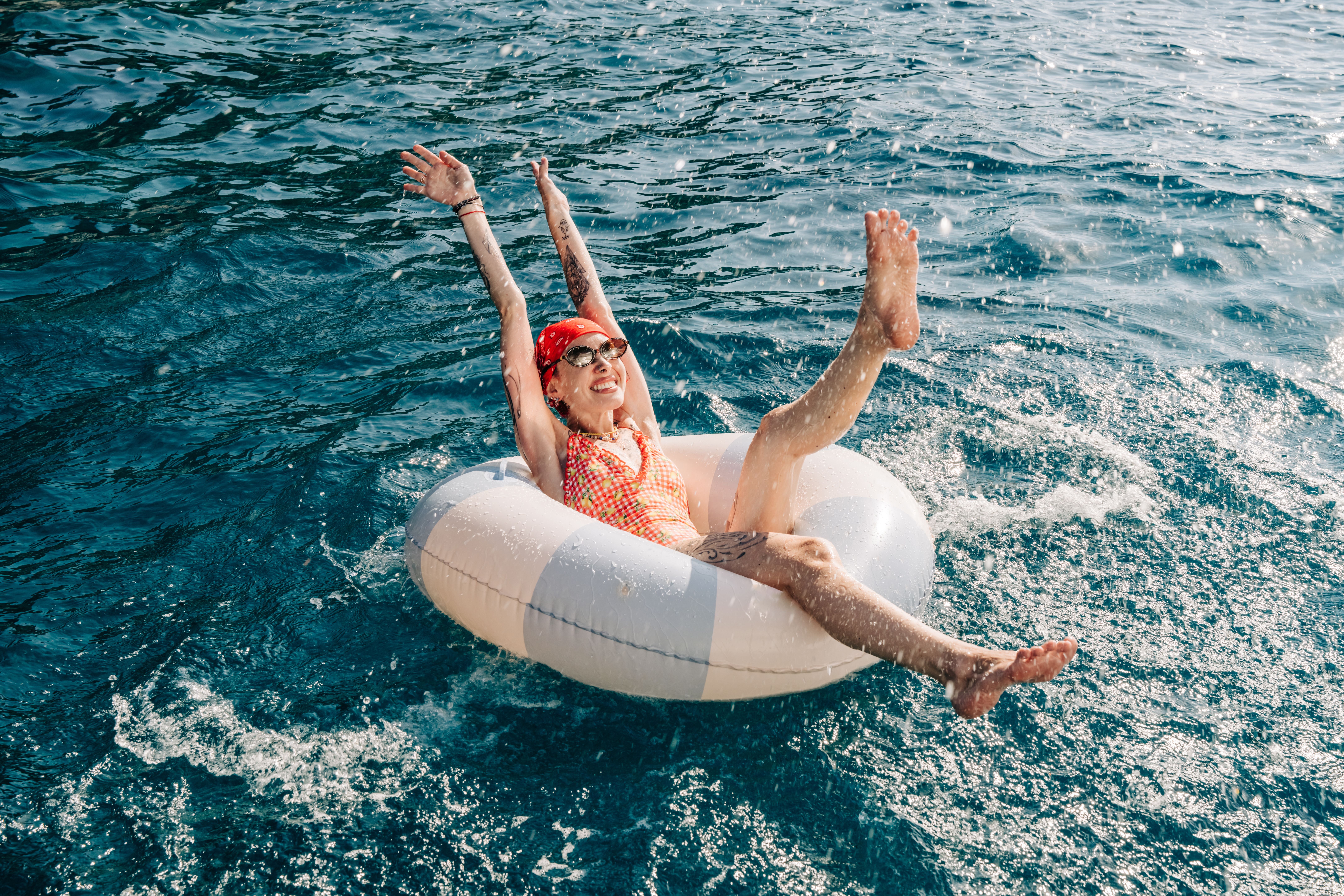 Frau mit Sonnenbrille und Schwimmring lacht im Wasser und streckt Arme und Beine nach oben.