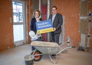 Zwei Personen stehen in einem Rohbau und halten ein Schild mit der Aufschrift „Bauen + Wohnen“.
