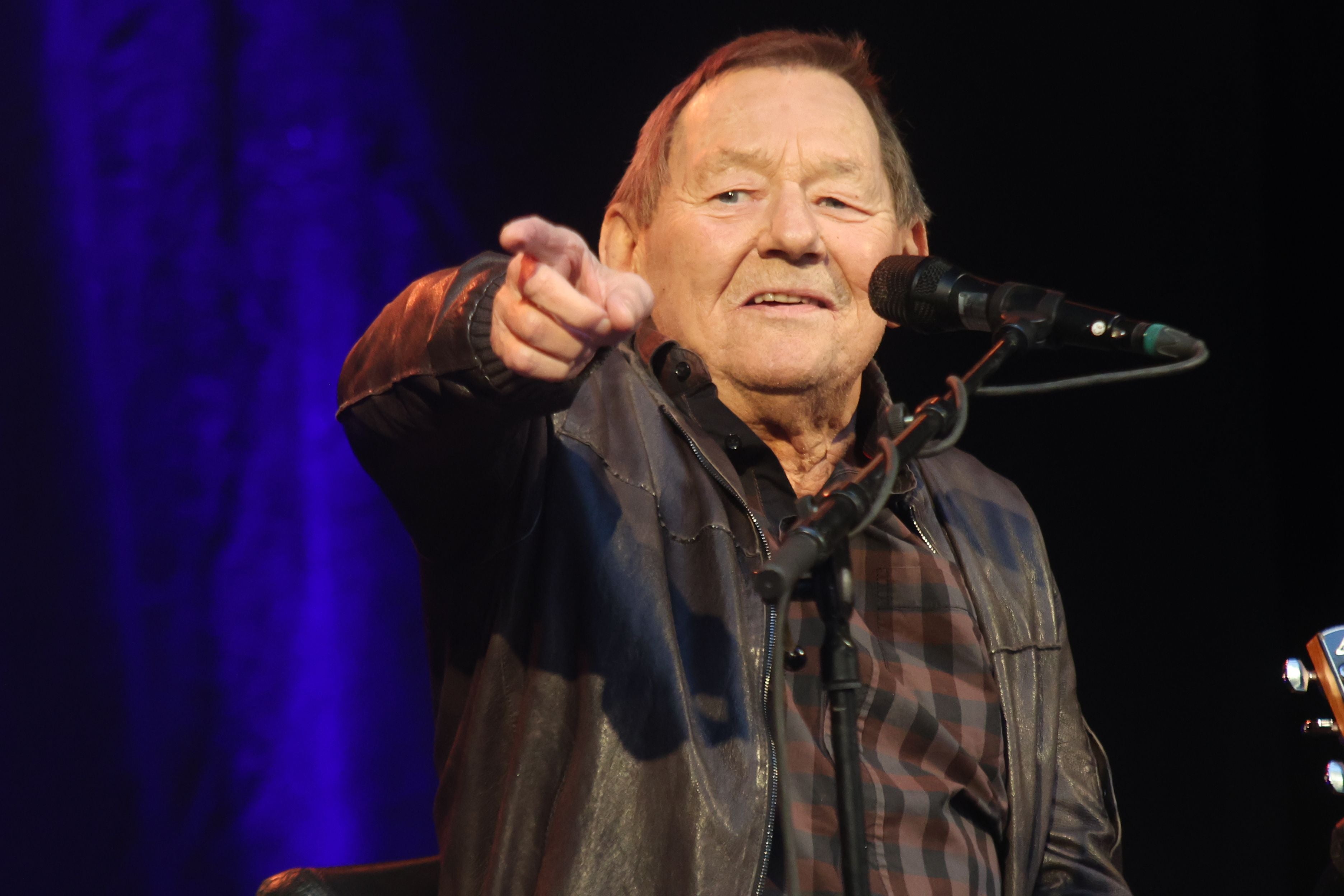 Ein älterer Mann singt auf der Bühne und zeigt lächelnd in die Kamera.