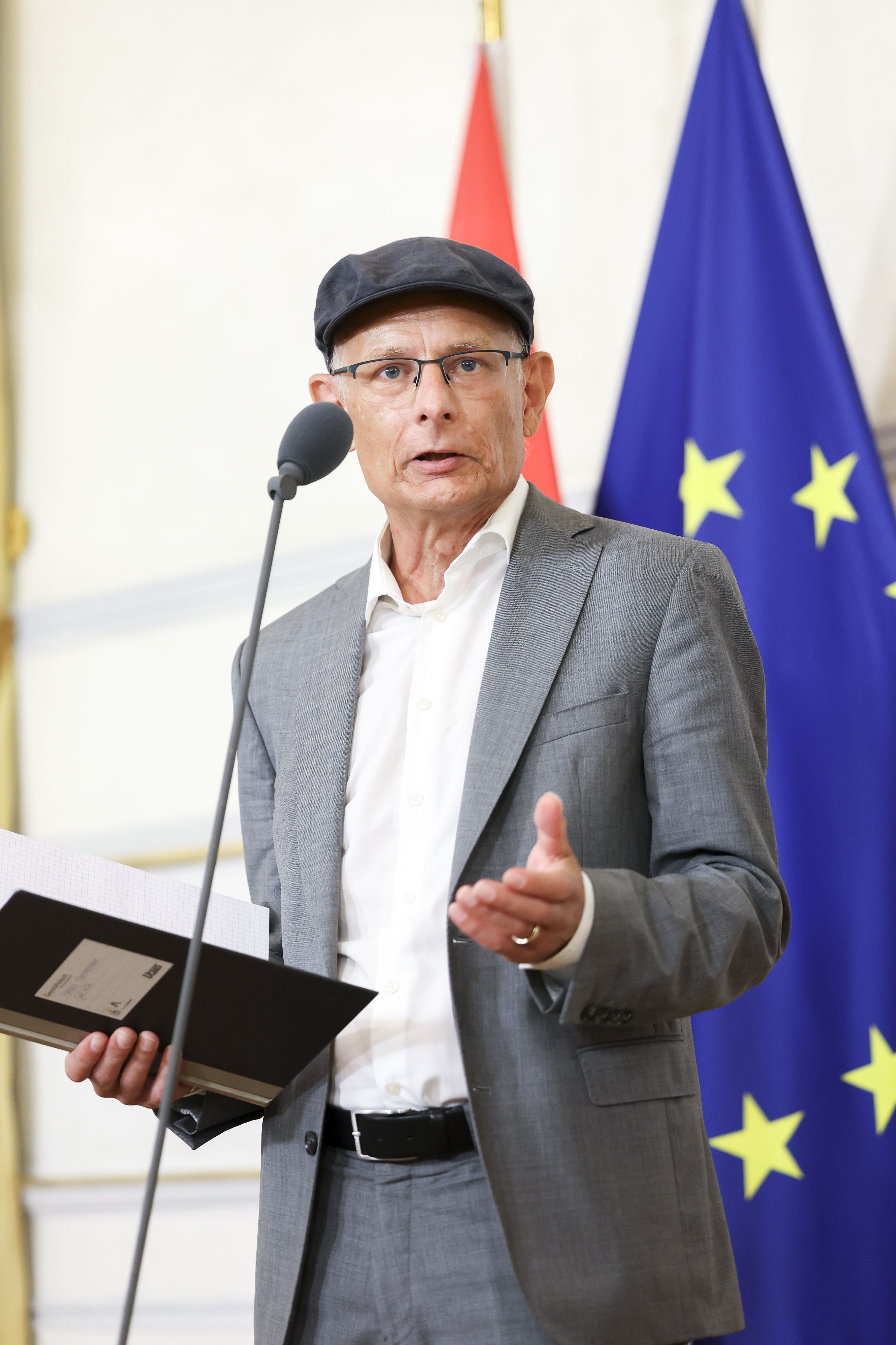 Ein Mann im grauen Anzug und Schirmmütze spricht bei einer Pressekonferenz vor EU-Flagge.