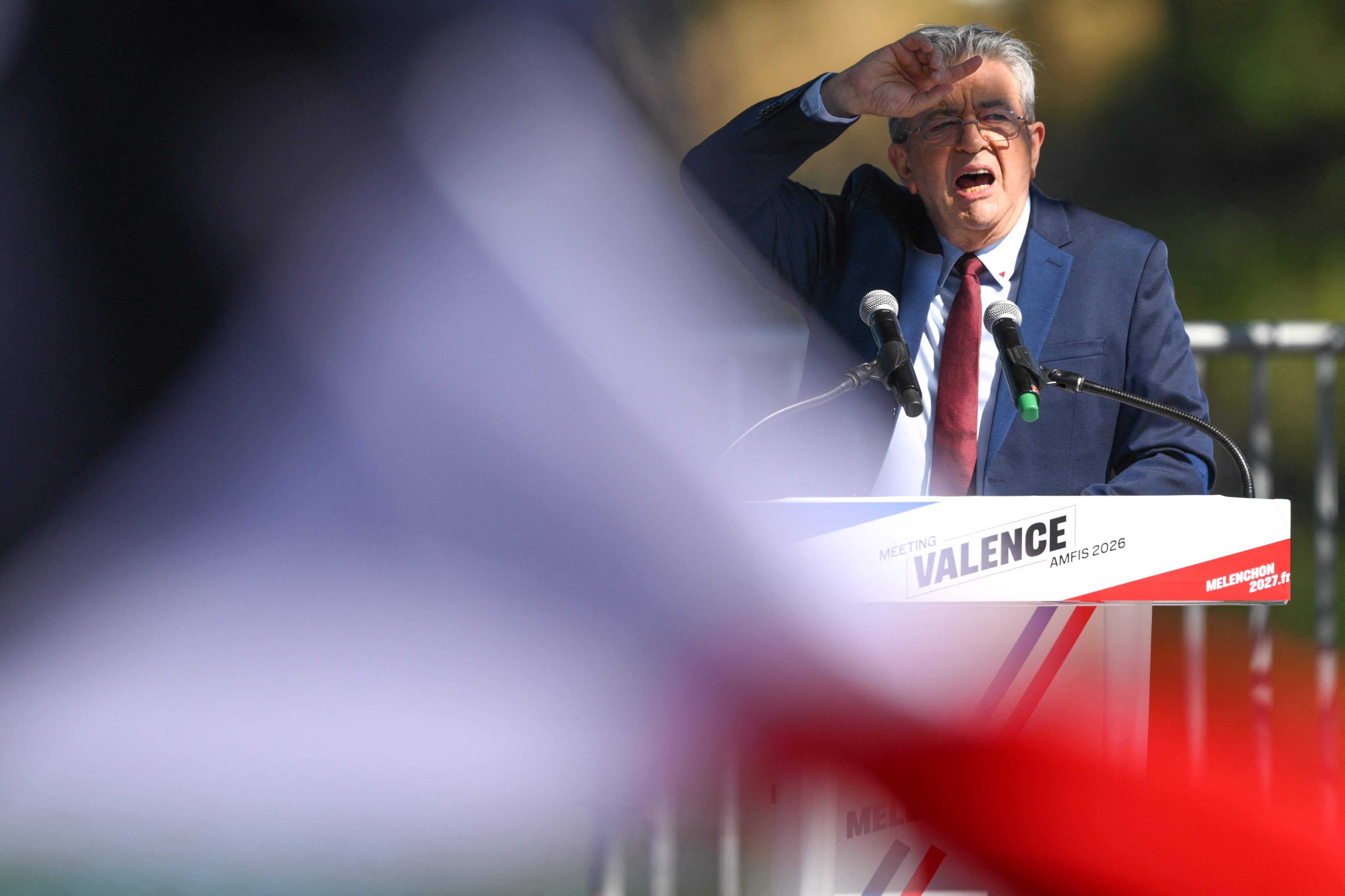 Ein Mann hält eine Rede an einem Rednerpult, Vordergrund unscharfe Farben einer Flagge.