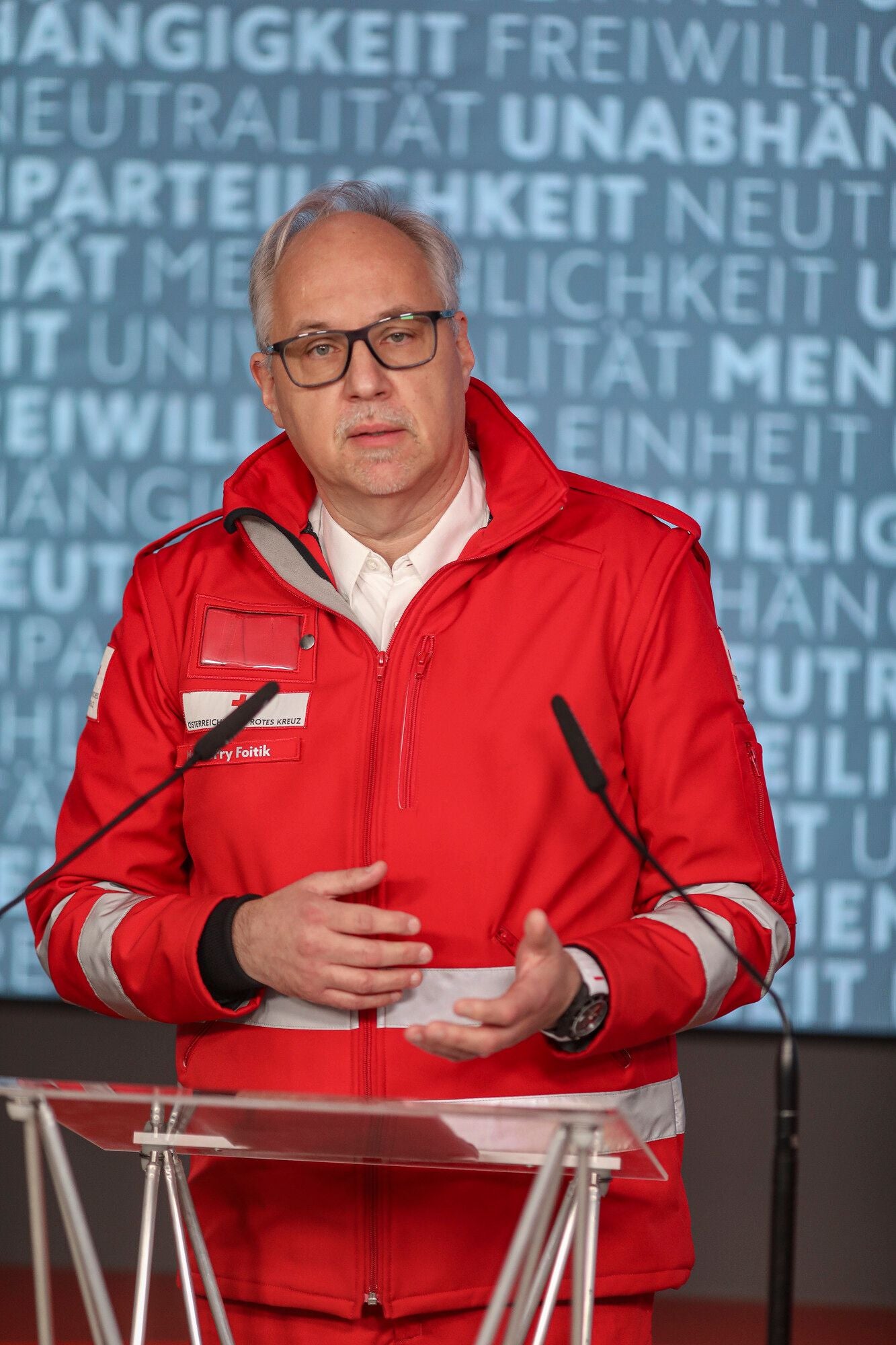 Ein Mann in roter Einsatzjacke spricht bei einer Pressekonferenz des Roten Kreuzes am Rednerpult.