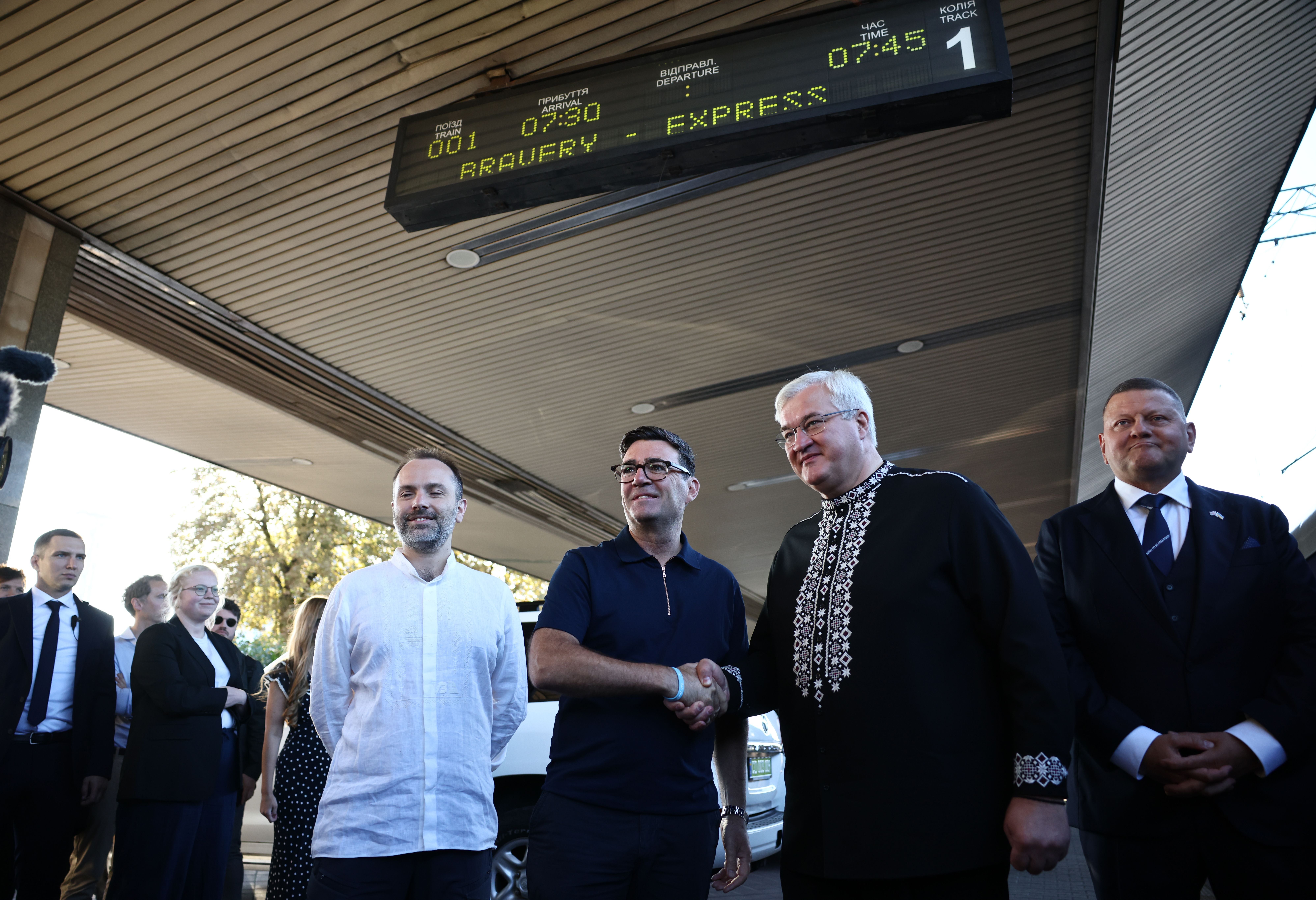 Mehrere Männer in Anzügen begrüßen sich unter einem Bahnhofsschild für den "Pravery Express".