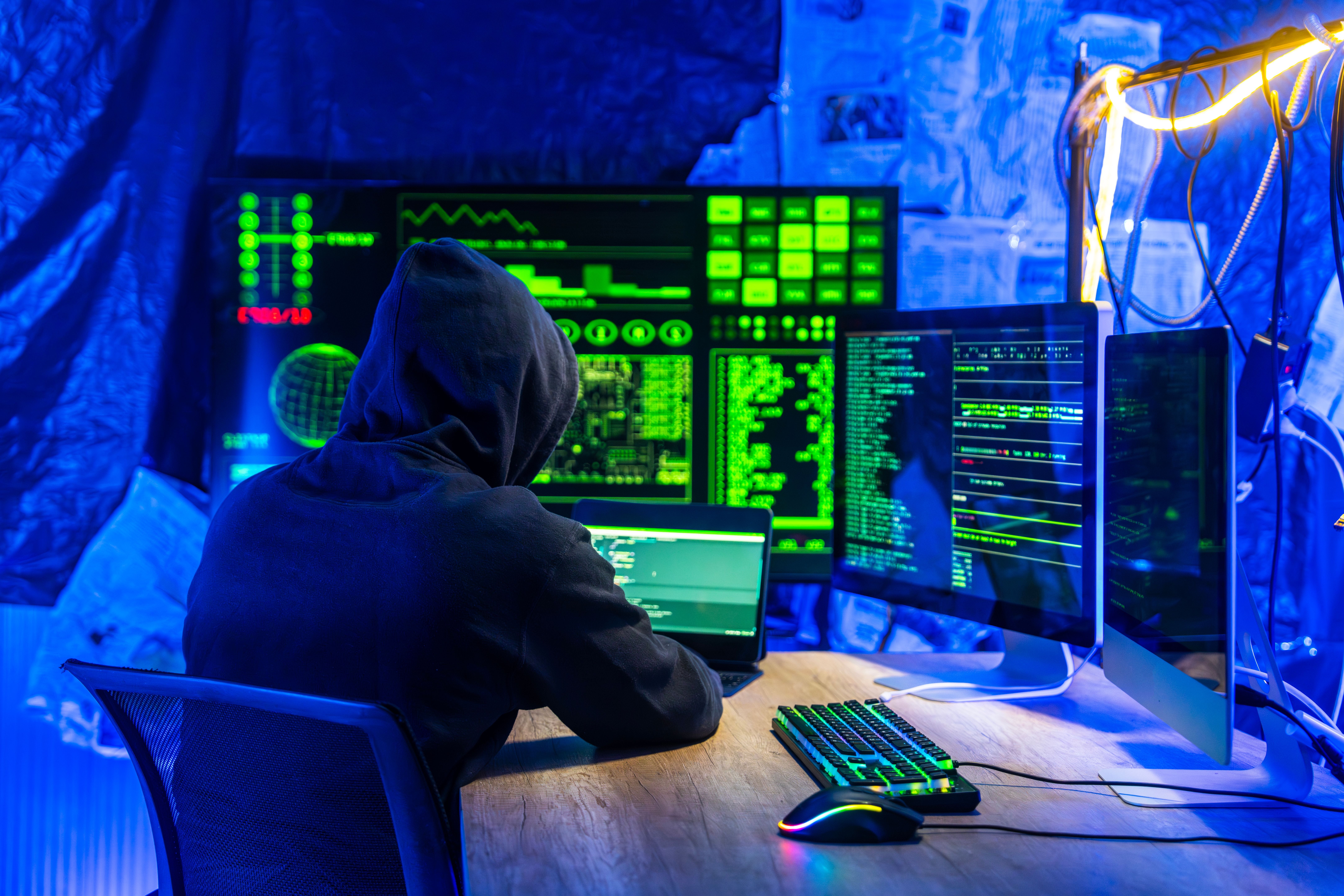 Person mit Kapuzenpullover arbeitet nachts an mehreren Monitoren mit grünem Code und Datenanzeigen.