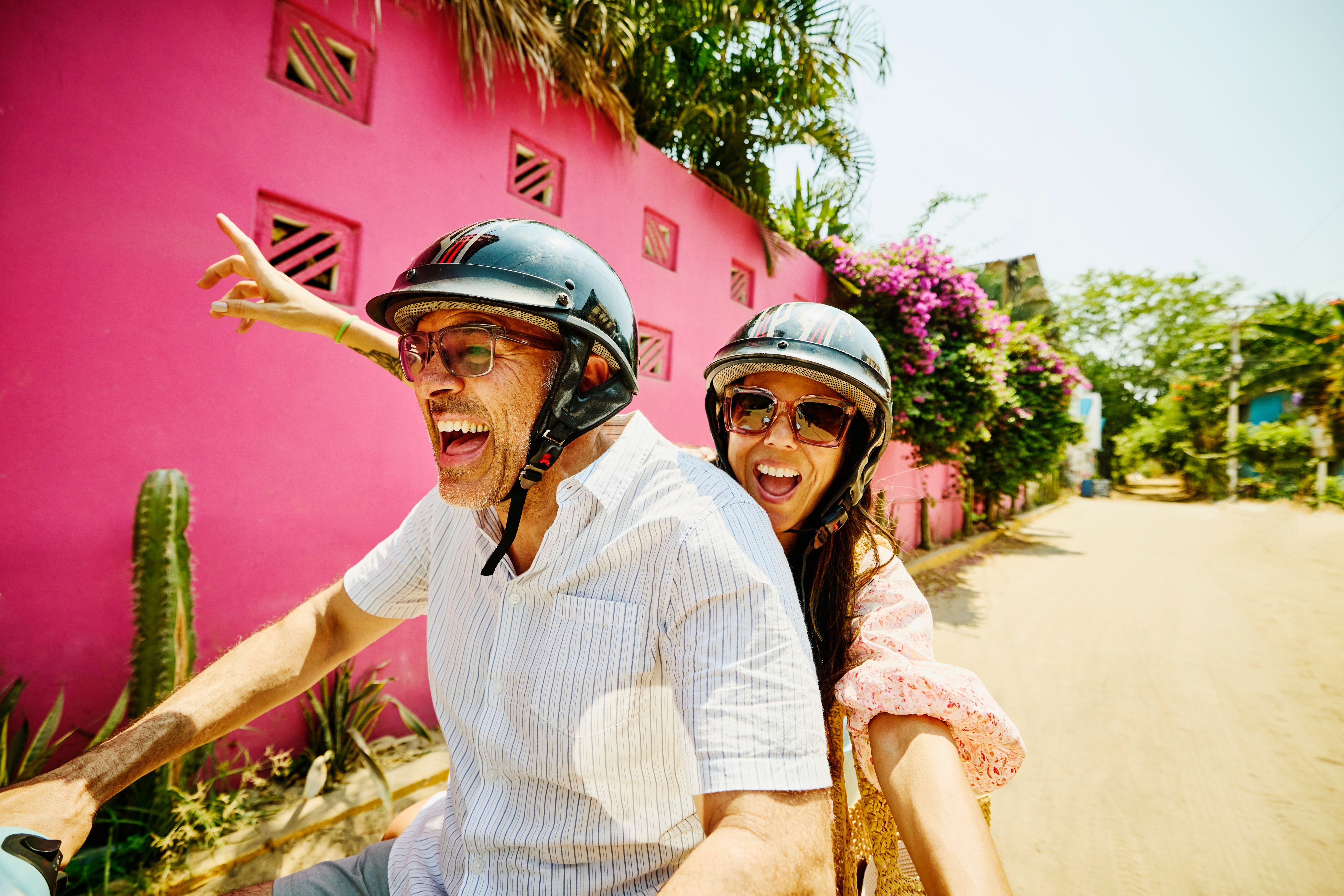 Fröhliches Paar mit Helmen fährt Moped auf leerer Straße vor pinker Wand im Urlaub.