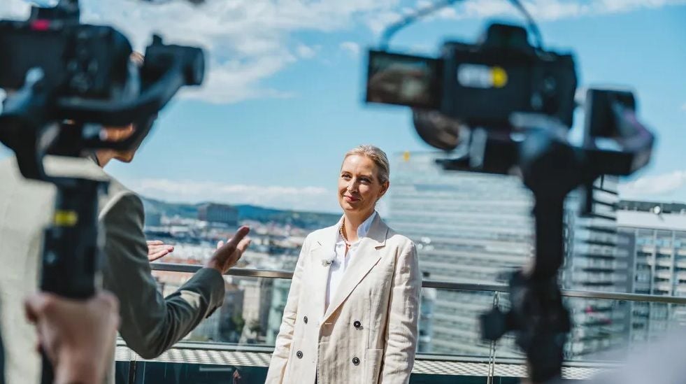 Frau im hellen Blazer wird auf einer Dachterrasse von Kameras gefilmt und interviewt.
