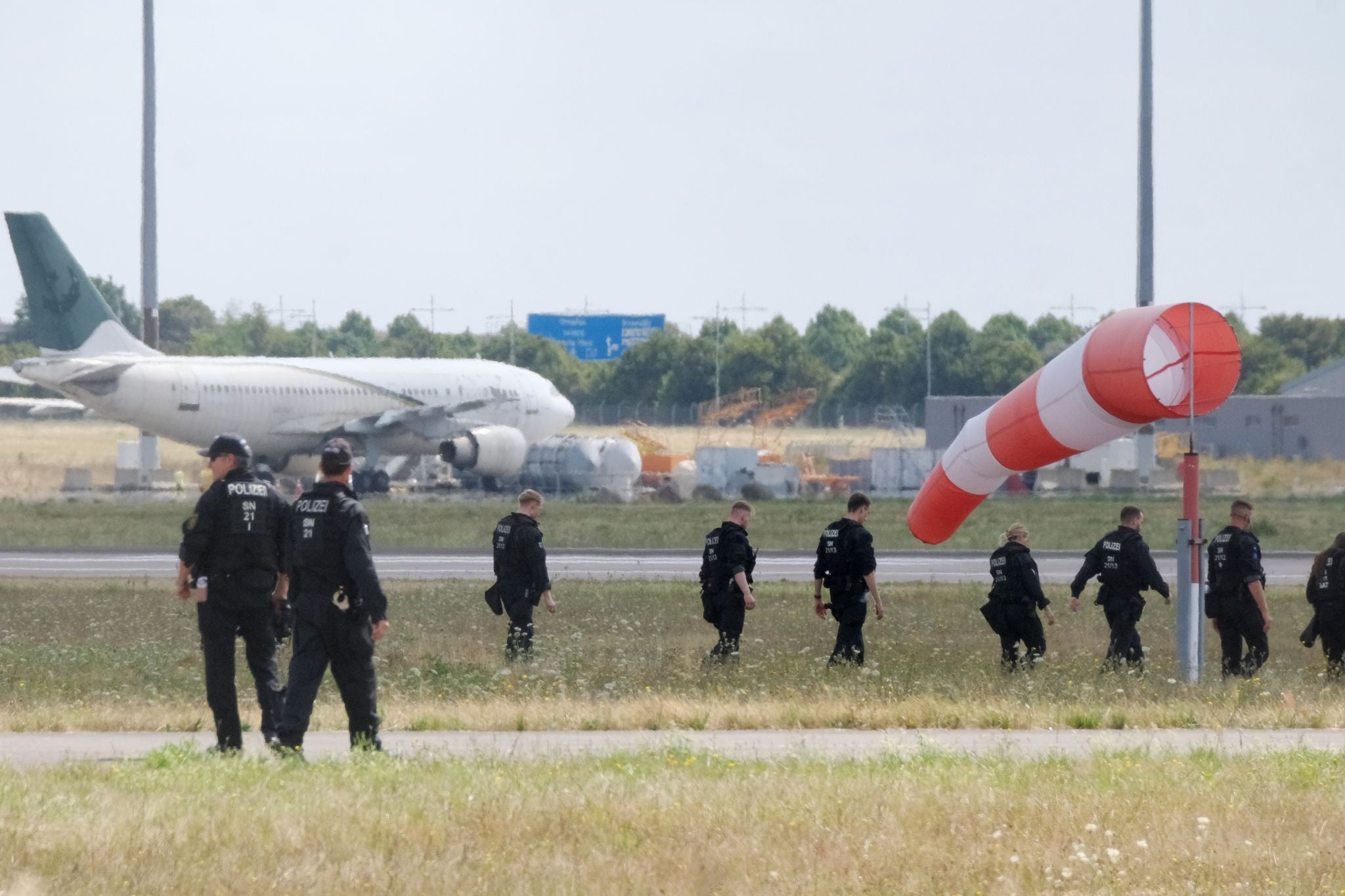 Polizisten durchsuchen zu Fuß eine Wiese am Flughafen Leipzig, im Hintergrund ein Flugzeug und Windsack.