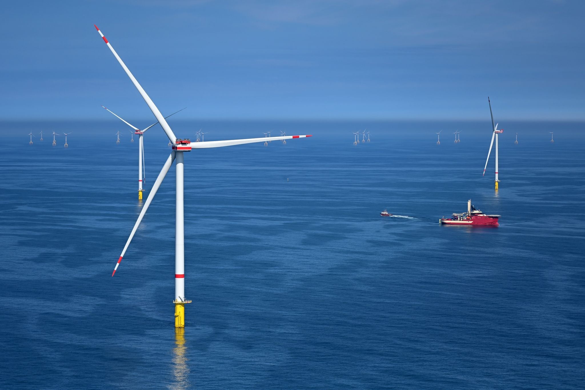 Offshore-Windkraftanlagen im Meer mit Wartungsschiff bei blauem Himmel und ruhigem Wasser.
