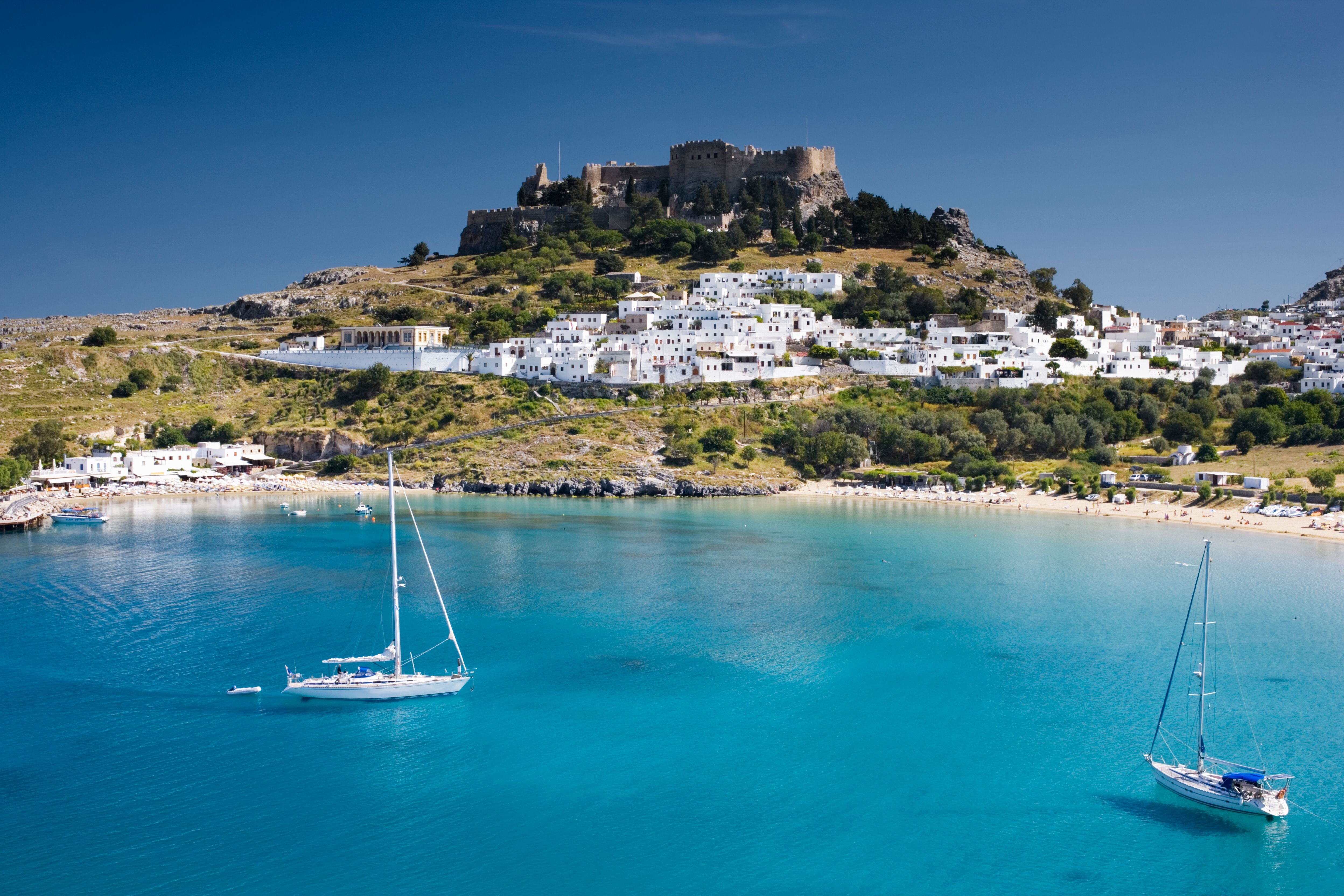 Blick auf eine Burg, weiße Häuser und Segelboote im türkisblauen Wasser einer Bucht.