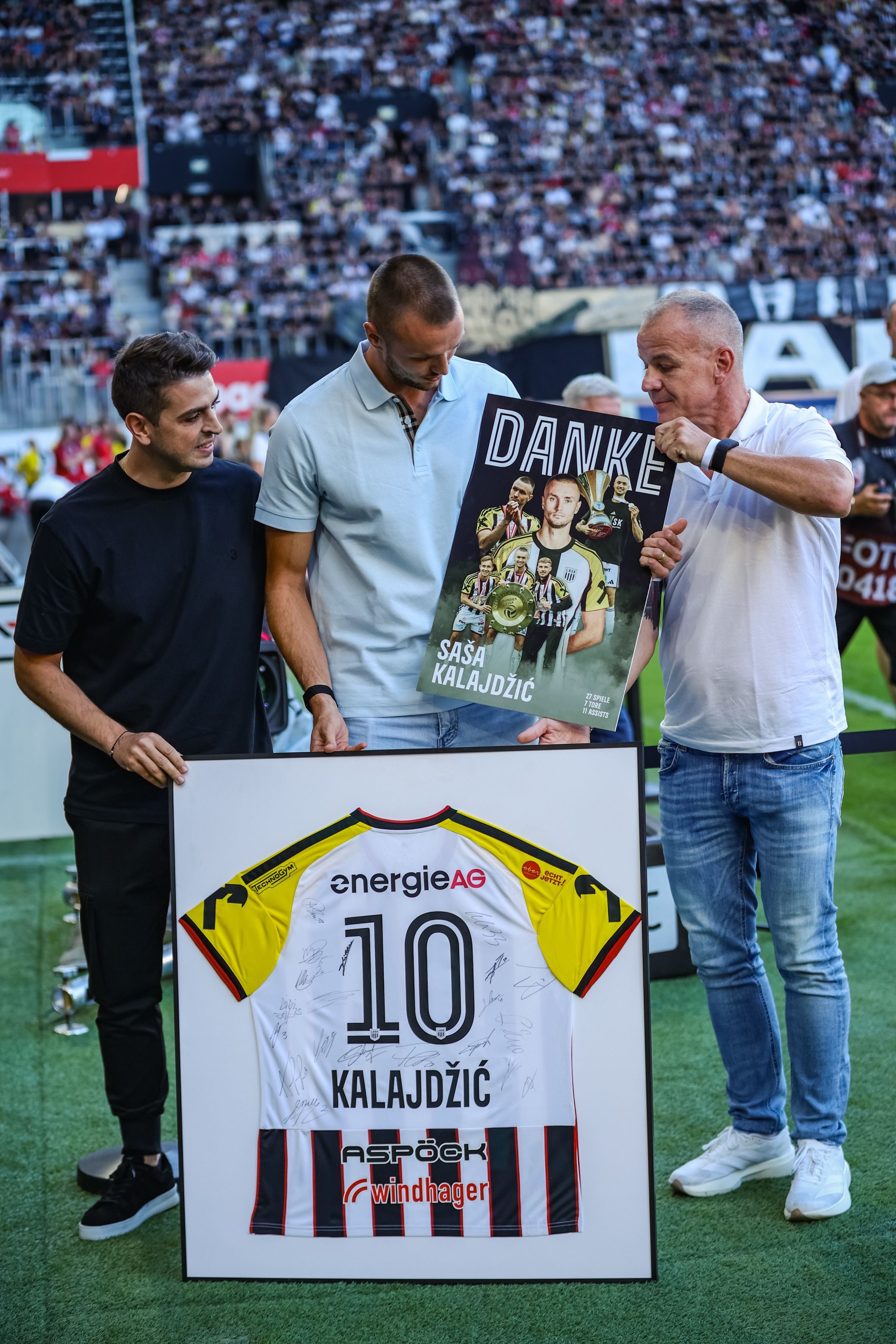 Drei Männer halten ein gerahmtes Trikot und ein Dankesplakat im Fußballstadion.