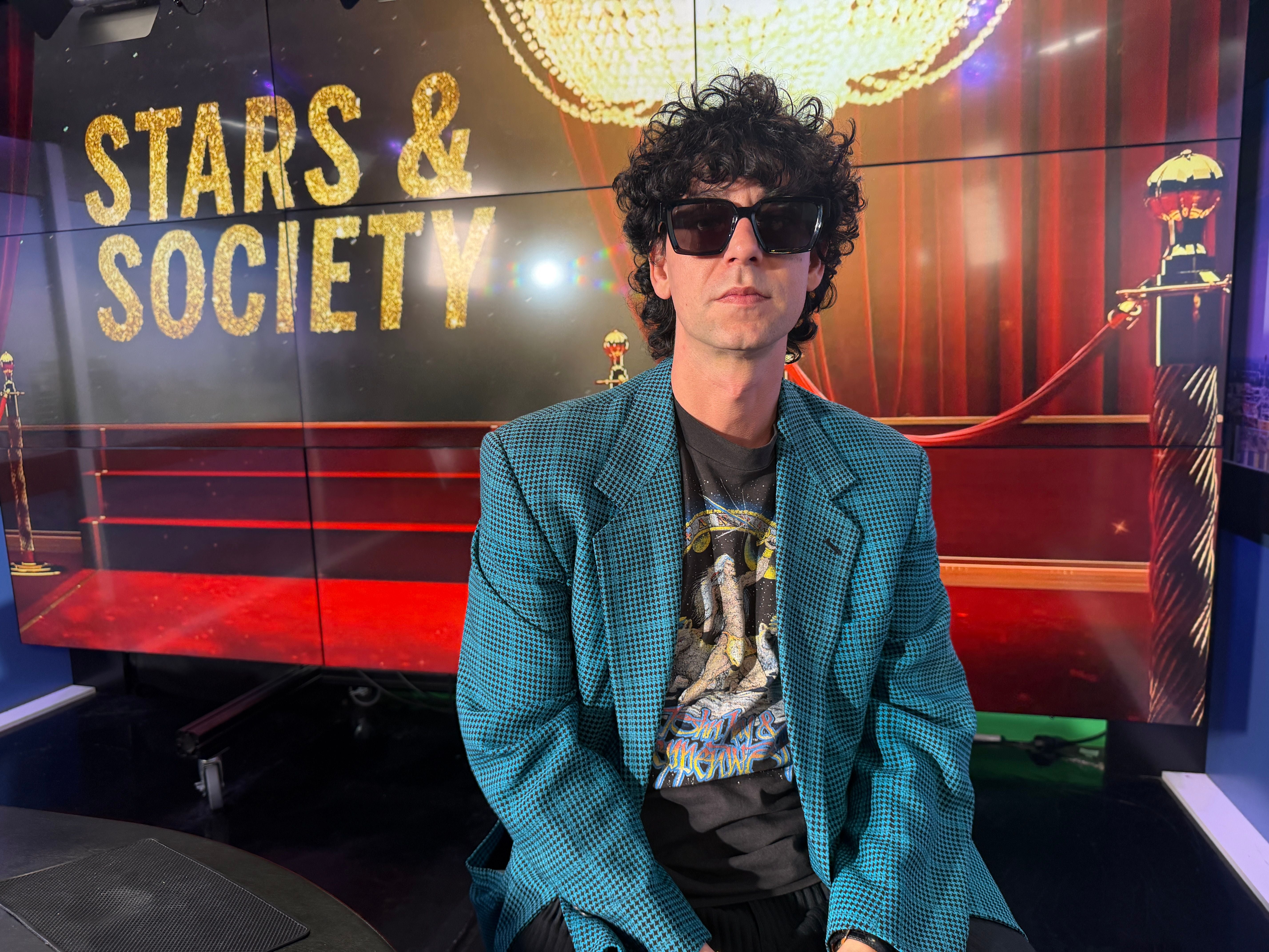 Ein Mann mit Sonnenbrille sitzt vor einer "Stars & Society"-Kulisse im Fernsehstudio.