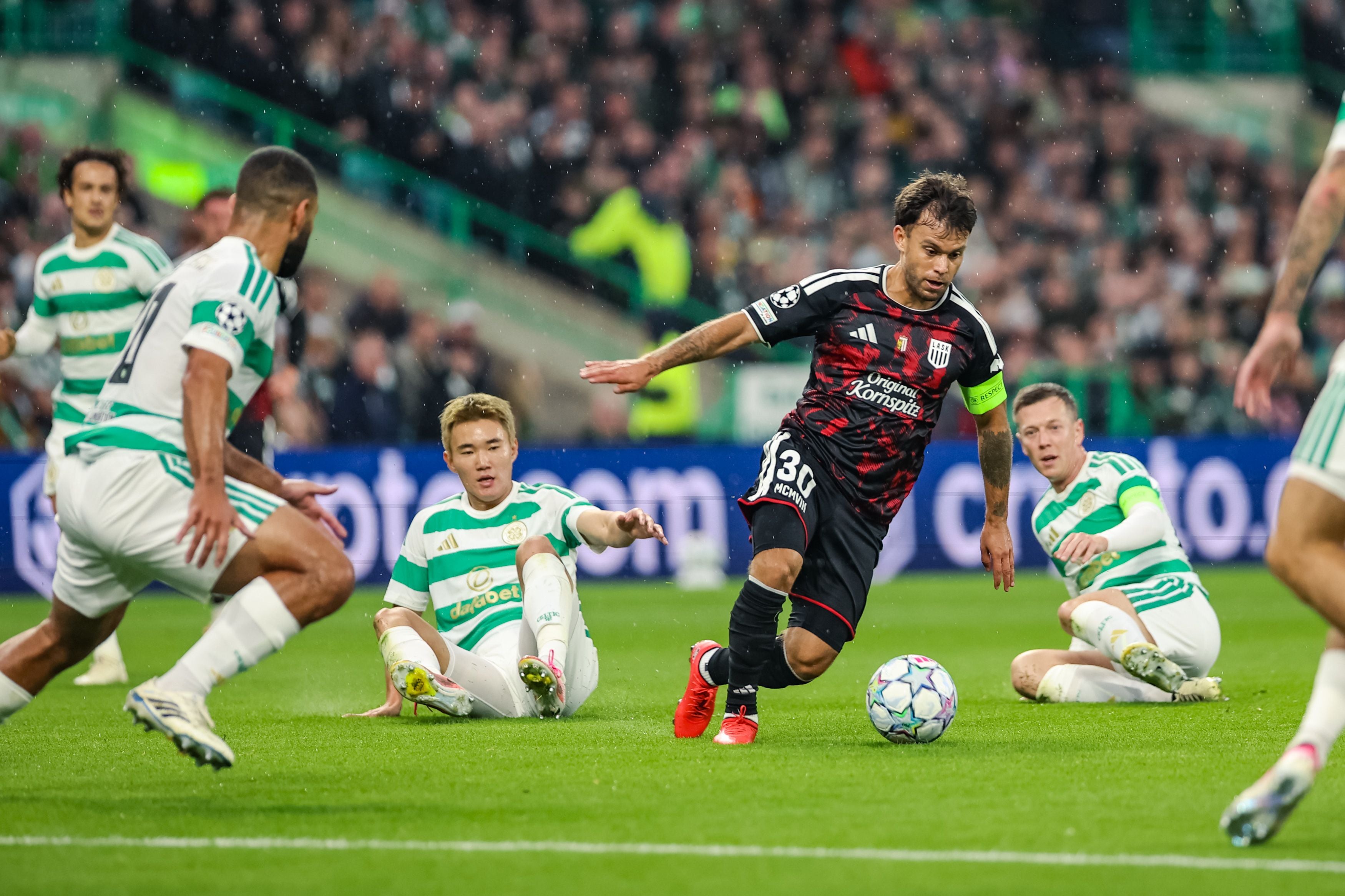LASK-Spieler&nbsp;Sasha Horvath in einem schwarz-roten Trikot dribbelt den Ball auf einem grünen Rasen, umgeben von Gegnern in weiß-grün gestreiften Trikots, während Zuschauer im Hintergrund auf den Tribünen sitzen.