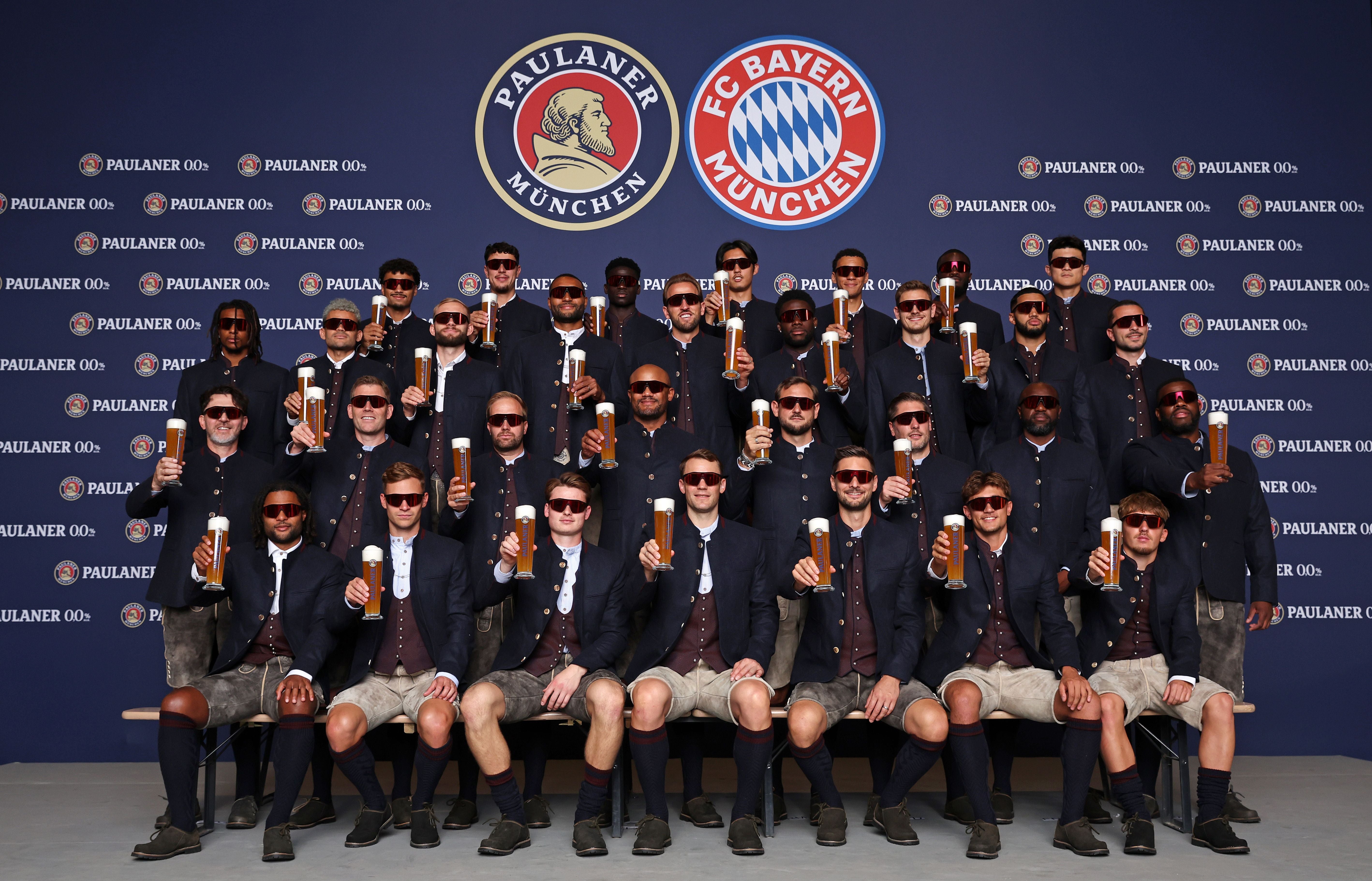 FC Bayern München Spieler posieren in Tracht mit Biergläsern vor Paulaner- und Vereinslogo.
