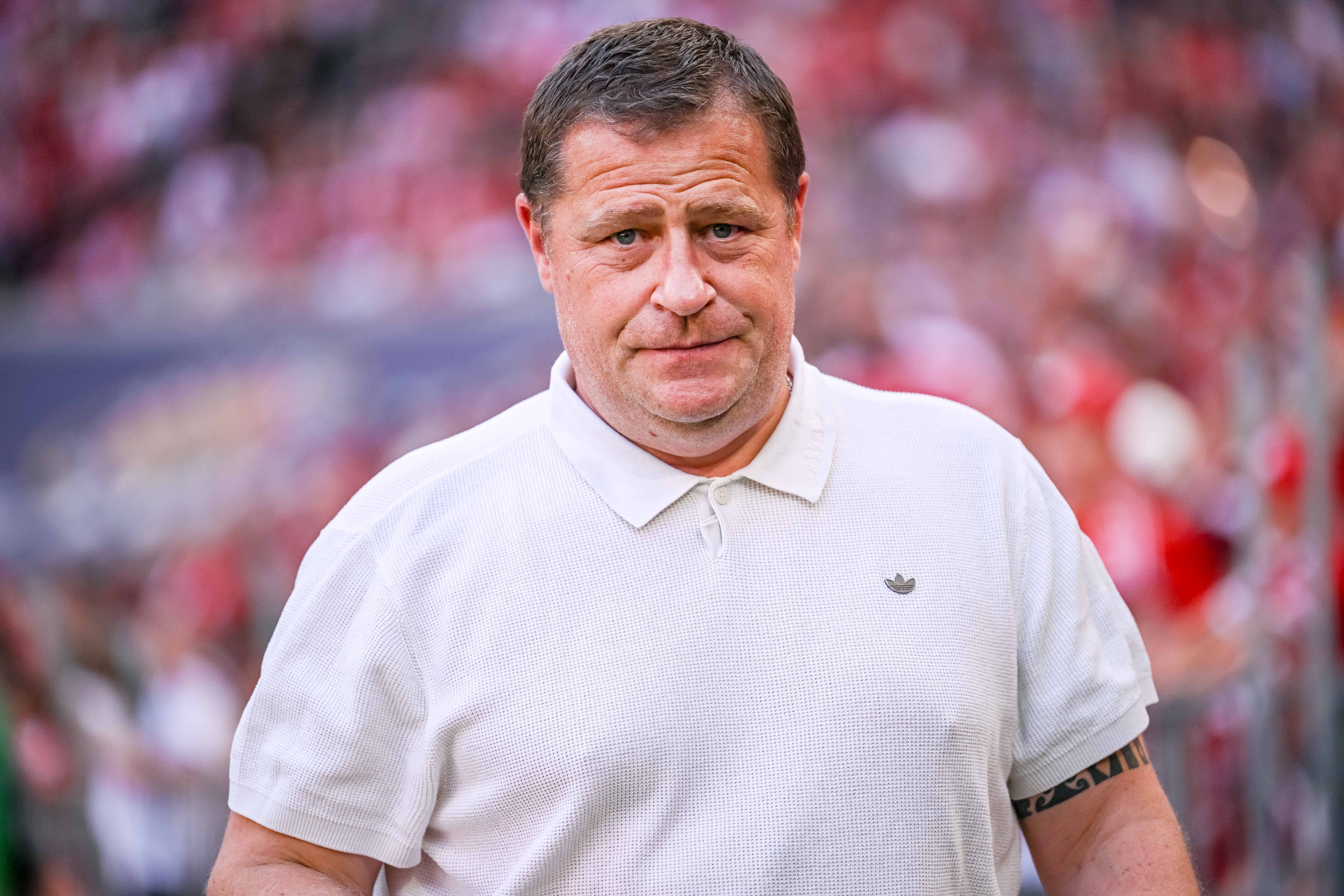 Ein Mann im weißen Poloshirt blickt ernst in die Kamera, unscharfer Stadionhintergrund.