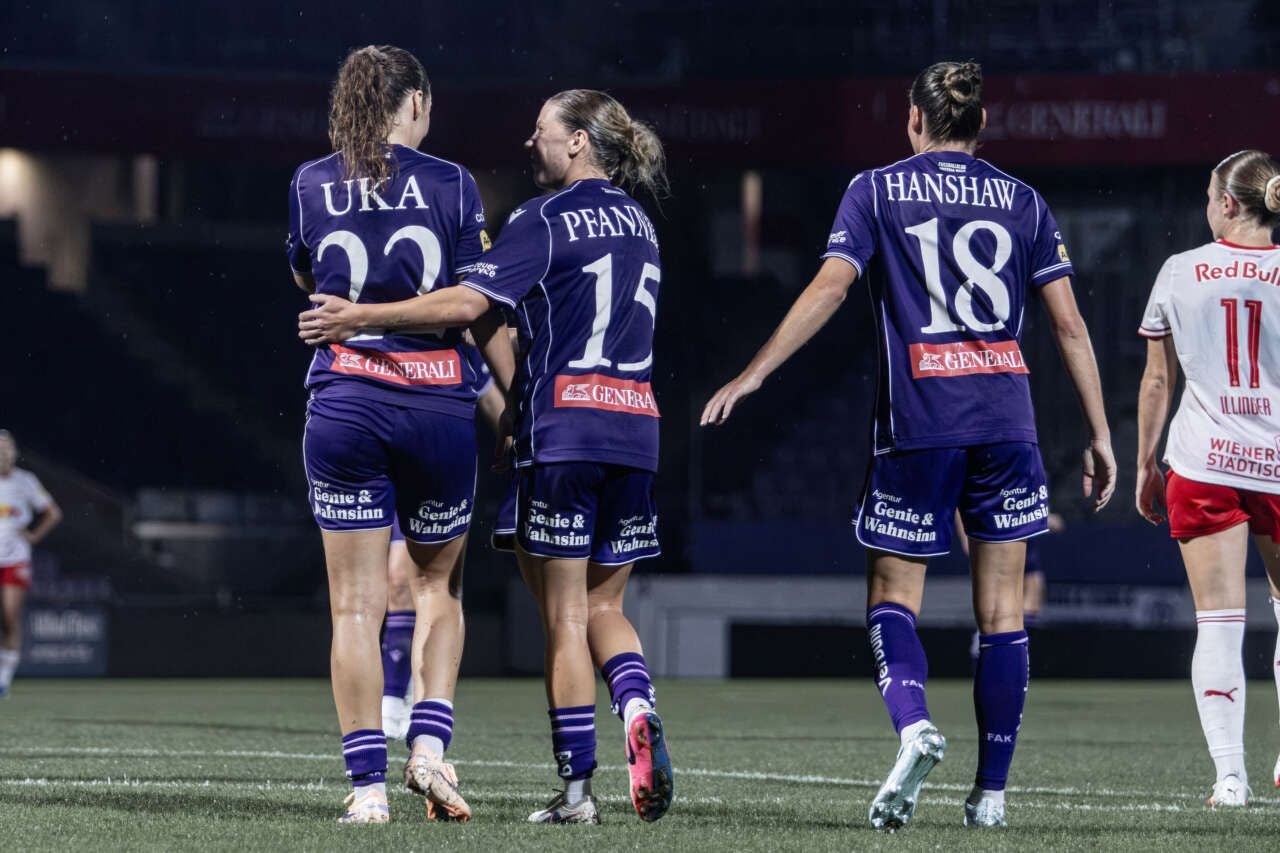 Drei Fußballspielerinnen im violetten Trikot mit den Aufschriften Uka, Pfattner und Hanshaw stehen von hinten gesehen auf dem Spielfeld. Eine von ihnen umfasst die andere an der Hüfte. Rechts ist eine gegnerische Spielerin im weiß-roten Trikot zu sehen.
