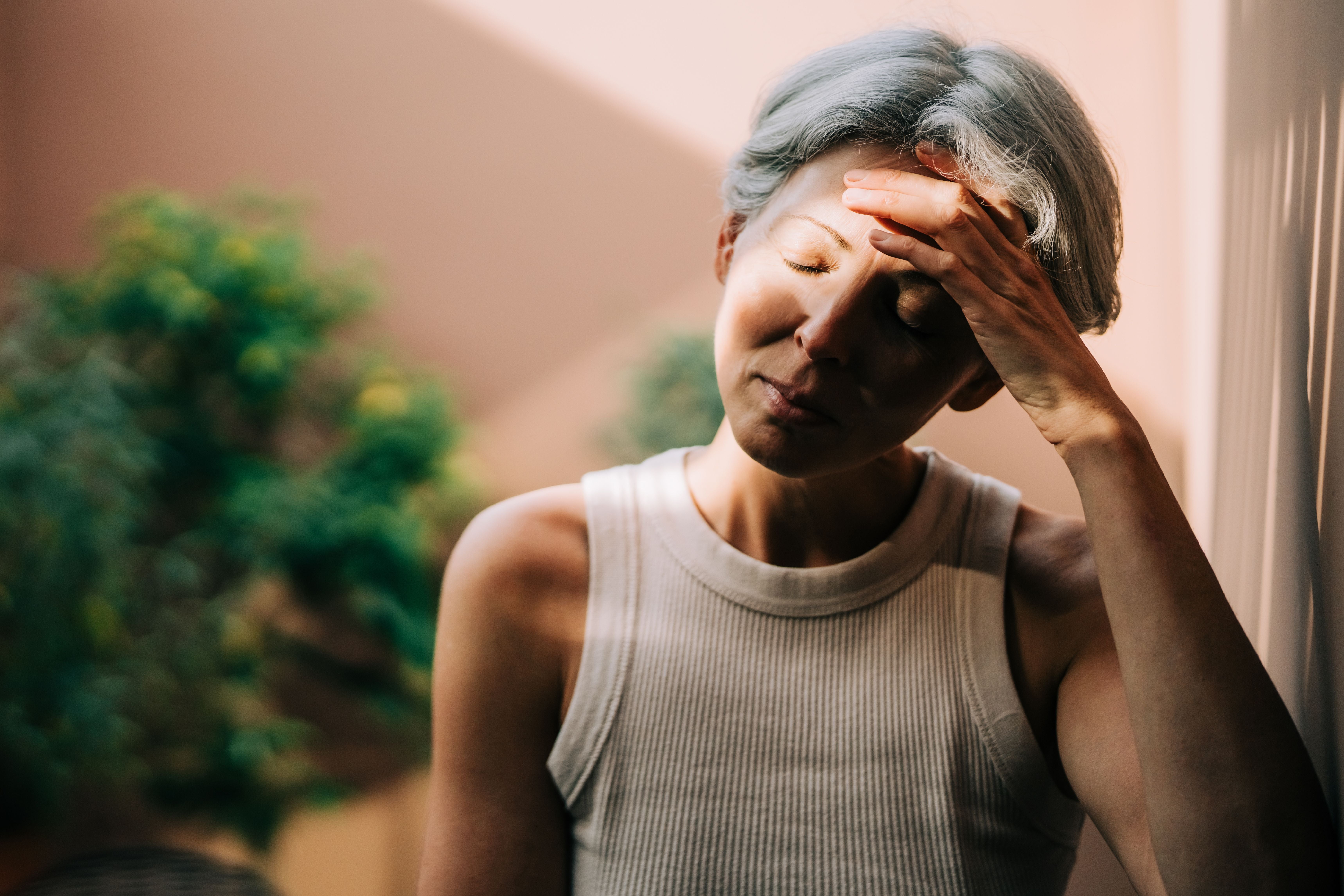 Frau mit grauen Haaren lehnt entspannt an einer Wand, Augen geschlossen, Hand an der Stirn im Sonnenlicht.