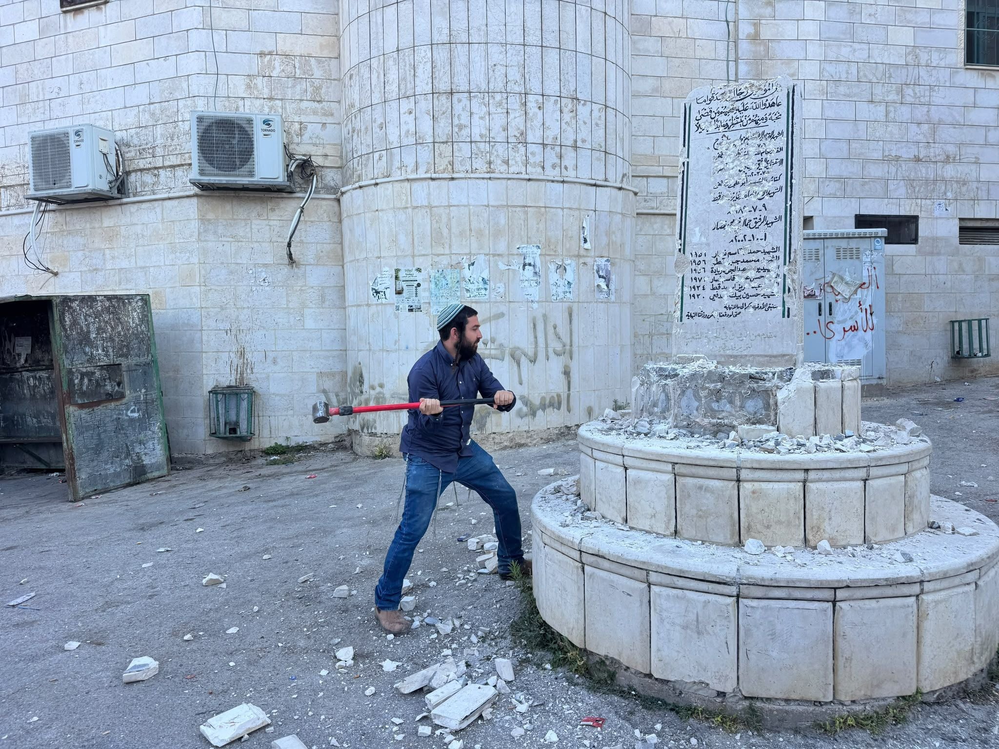 Ein Mann schlägt mit einem Vorschlaghammer auf ein beschädigtes Denkmal vor einem Gebäude ein.