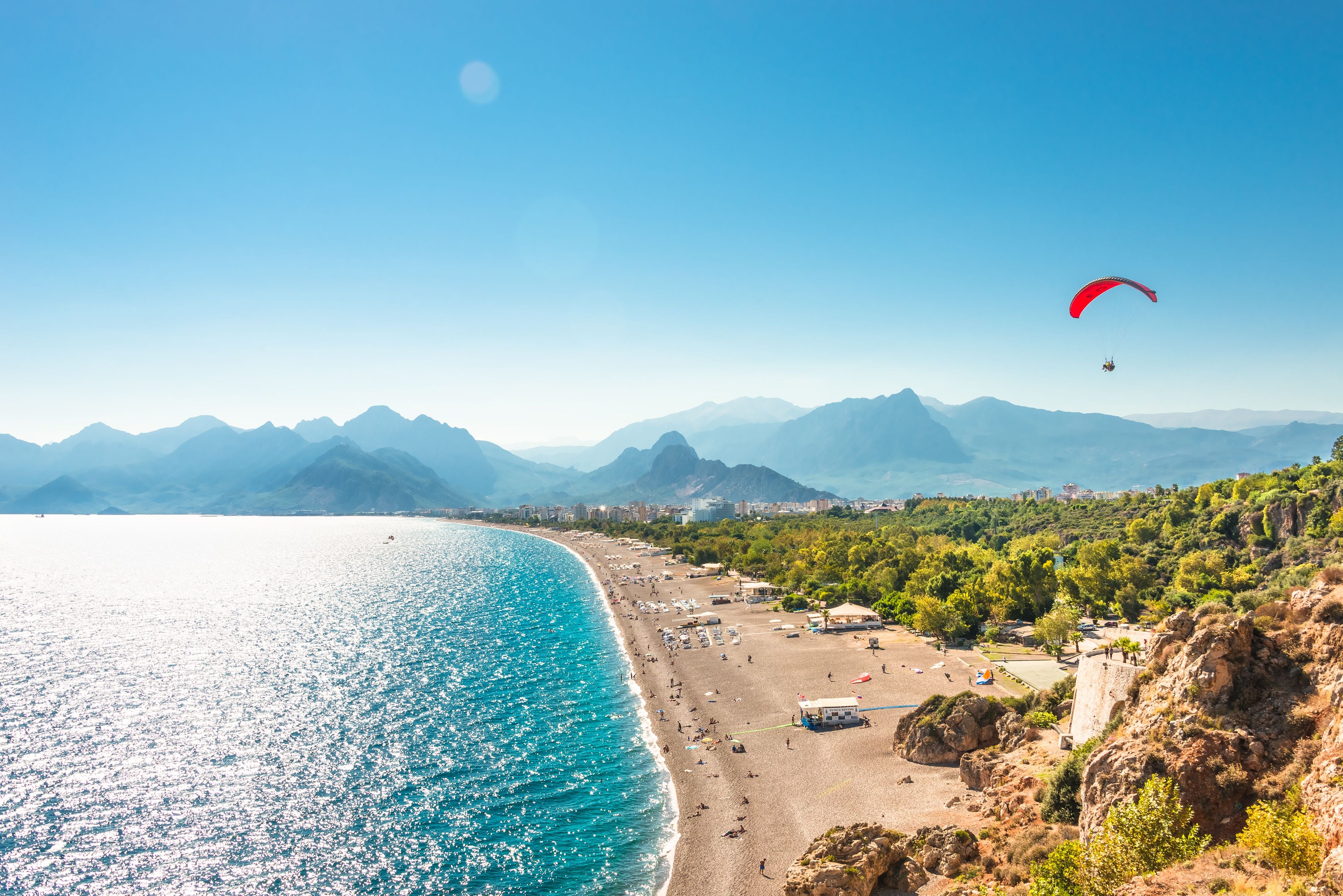 Panoramablick auf Antalyas Strand mit Gleitschirmflieger und Bergen im Hintergrund bei sonnigem Wetter.
