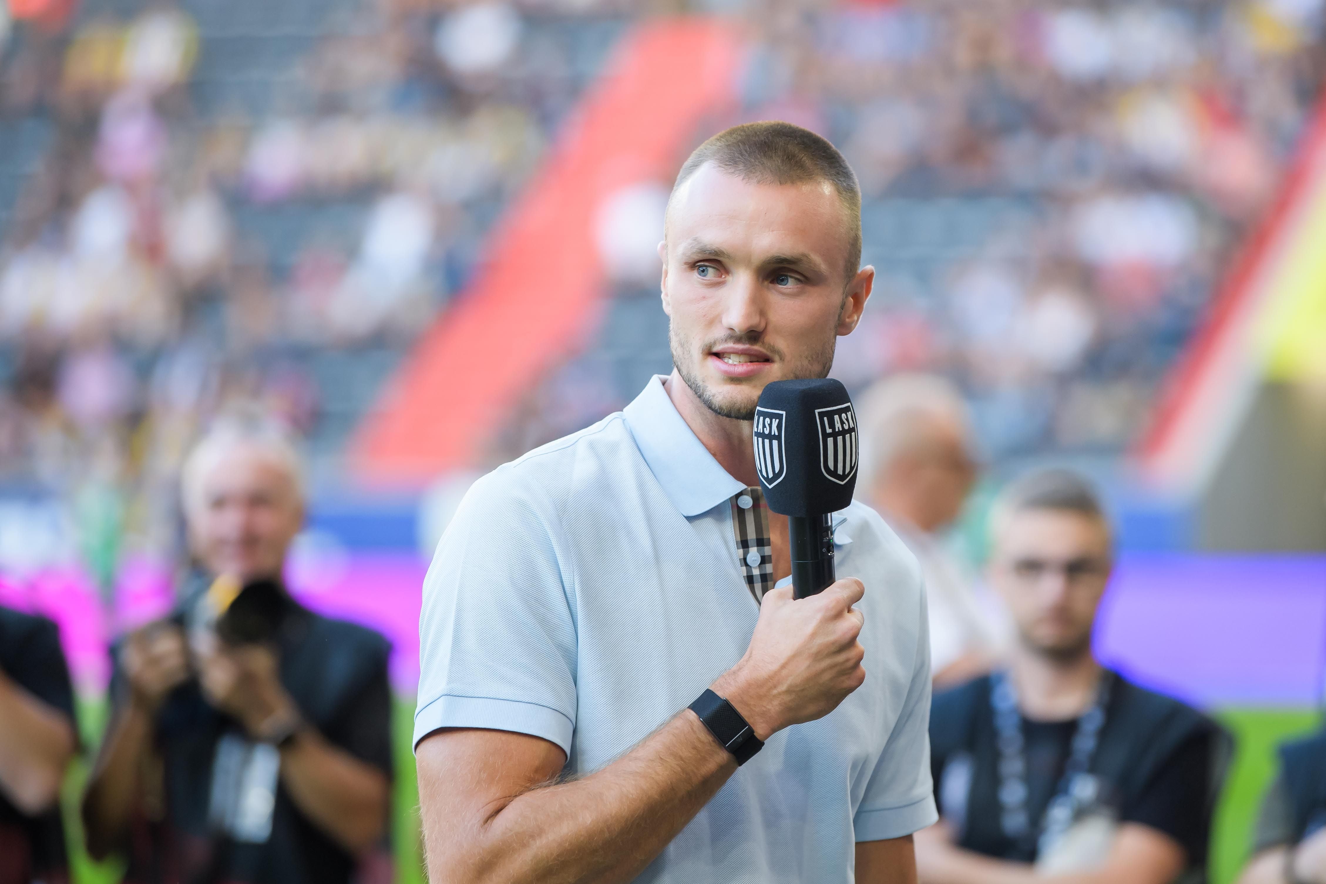 Ein Mann mit Mikrofon steht im Stadion und spricht, im Hintergrund unscharfe Zuschauer.