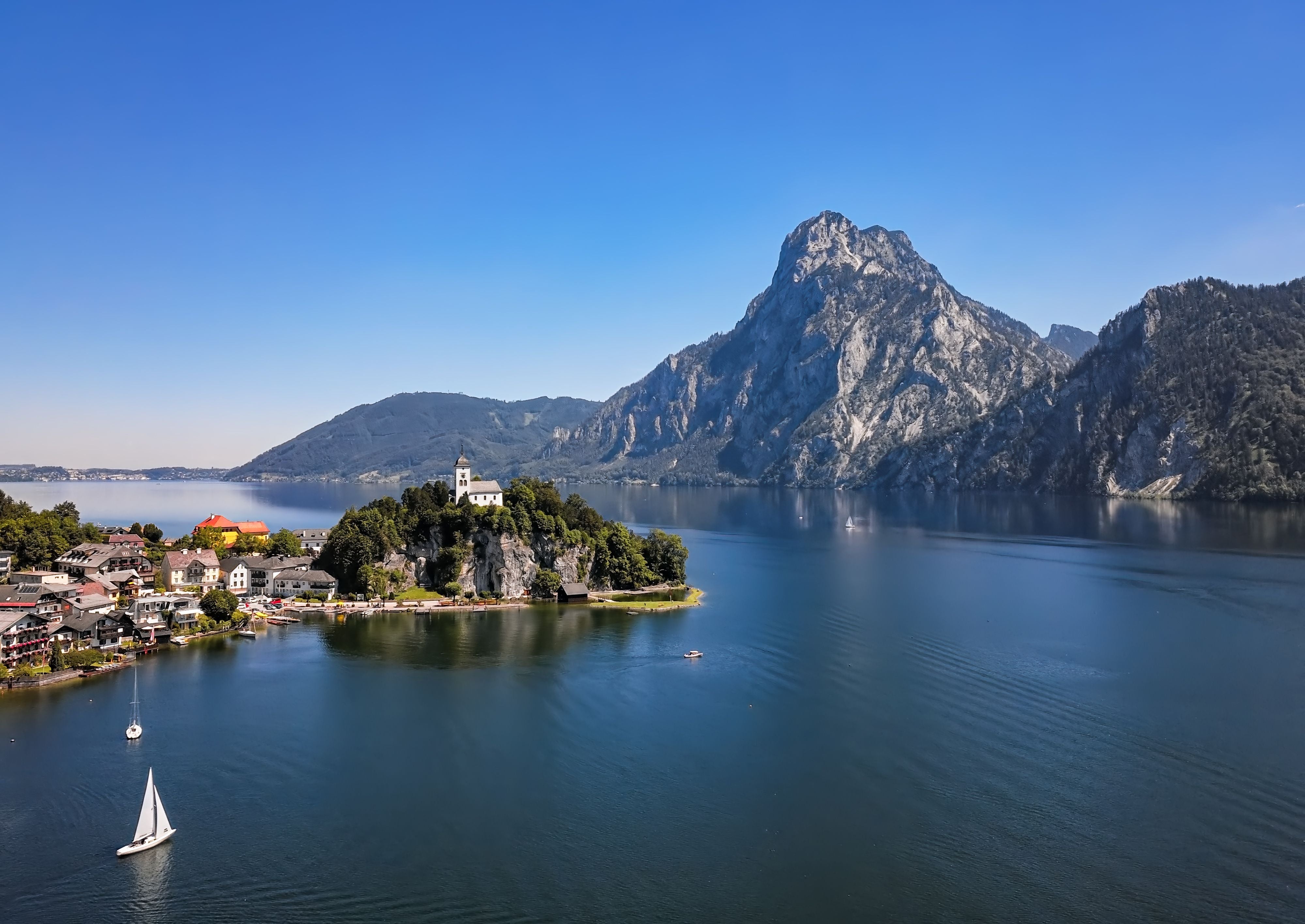 Alpensee mit Inselkirche, Segelbooten und hohen Bergen unter klarem, blauem Himmel bei Tageslicht.