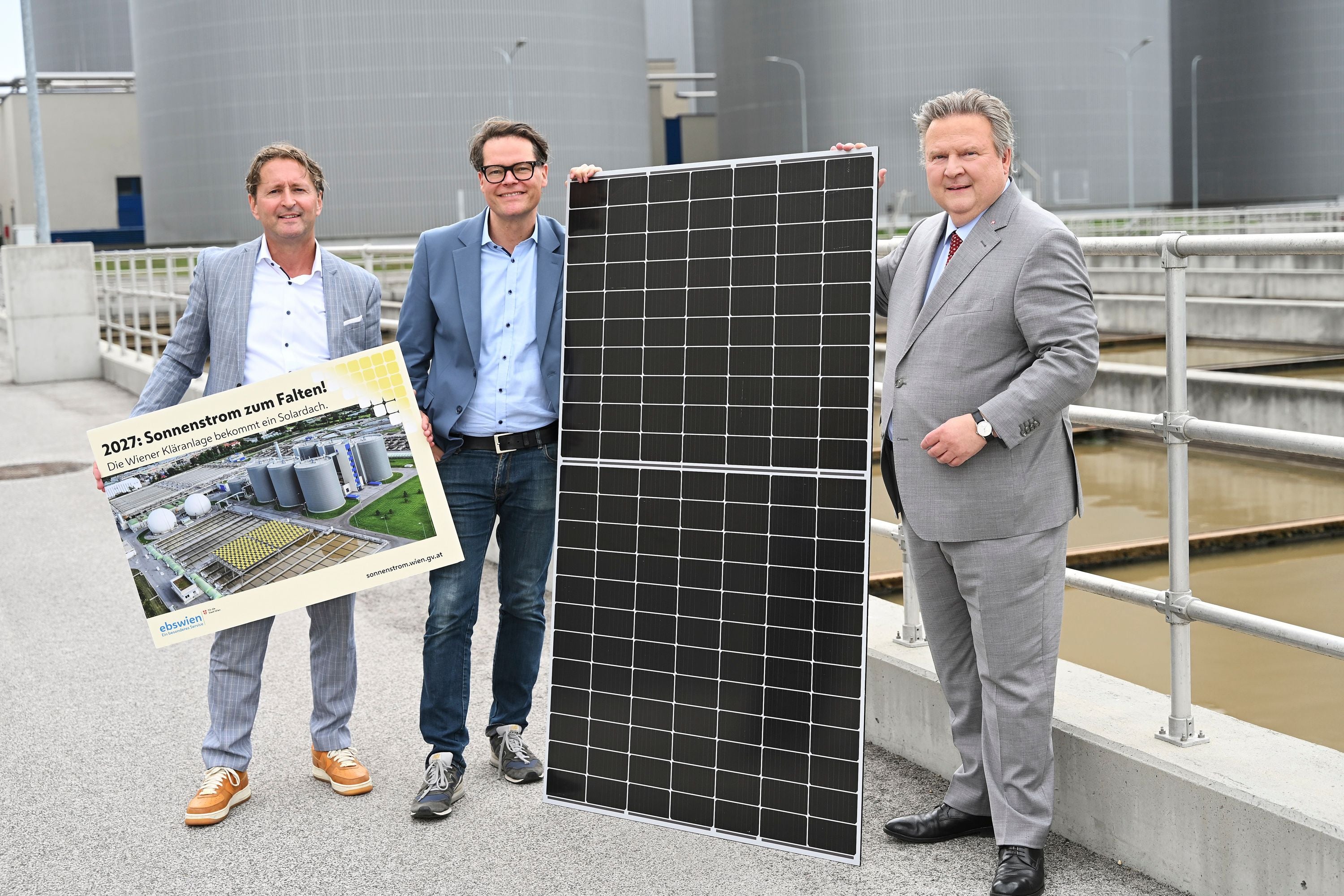 Drei Männer stehen vor einer Kläranlage, einer hält ein Solarpanel, ein anderer ein Plakat zur Solaranlage.