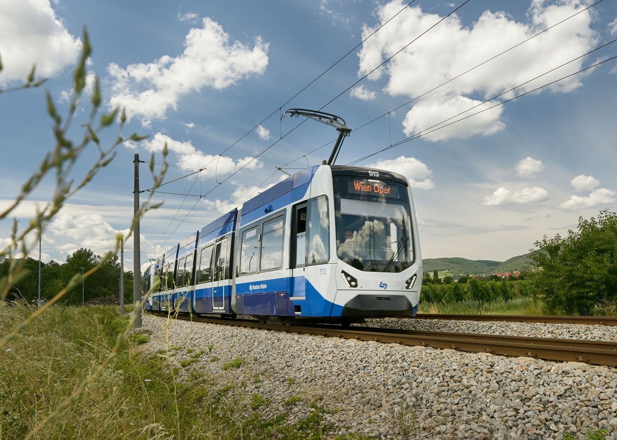 Eine blau-weiße Badner Bahn Straßenbahn fährt auf Gleisen durch eine ländliche Landschaft.