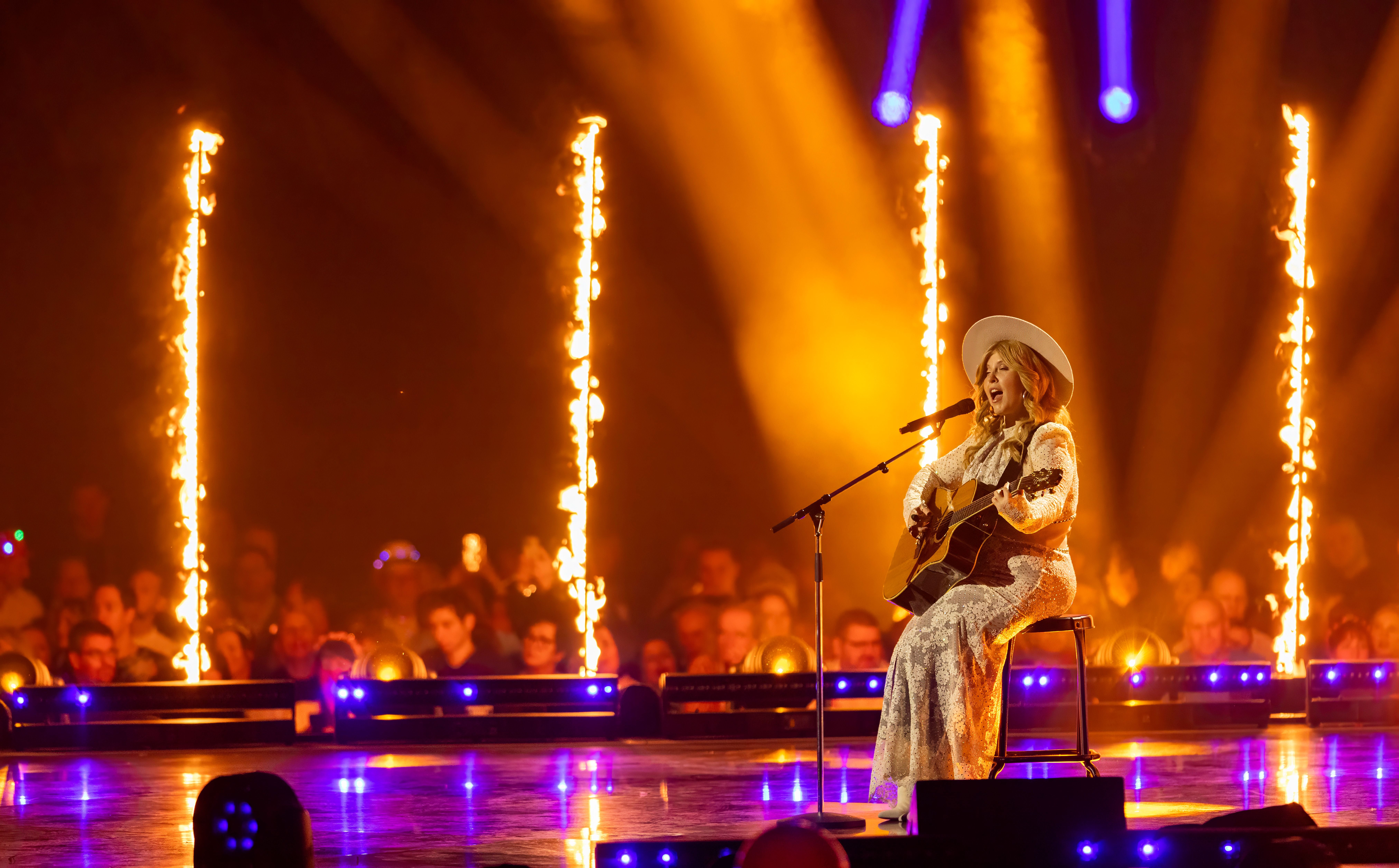 Eine Sängerin im weißen Kleid spielt Gitarre auf einer Bühne mit Flammensäulen und Publikum.