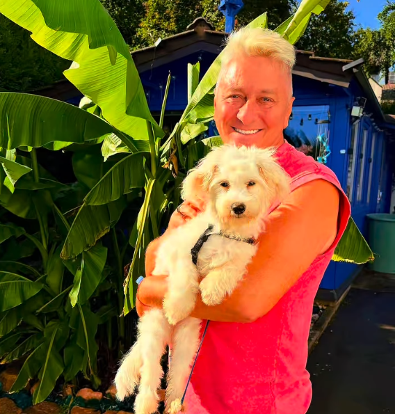 Mann im pinken Shirt hält einen kleinen weißen Hund im Arm, im Garten mit großen grünen Blättern.