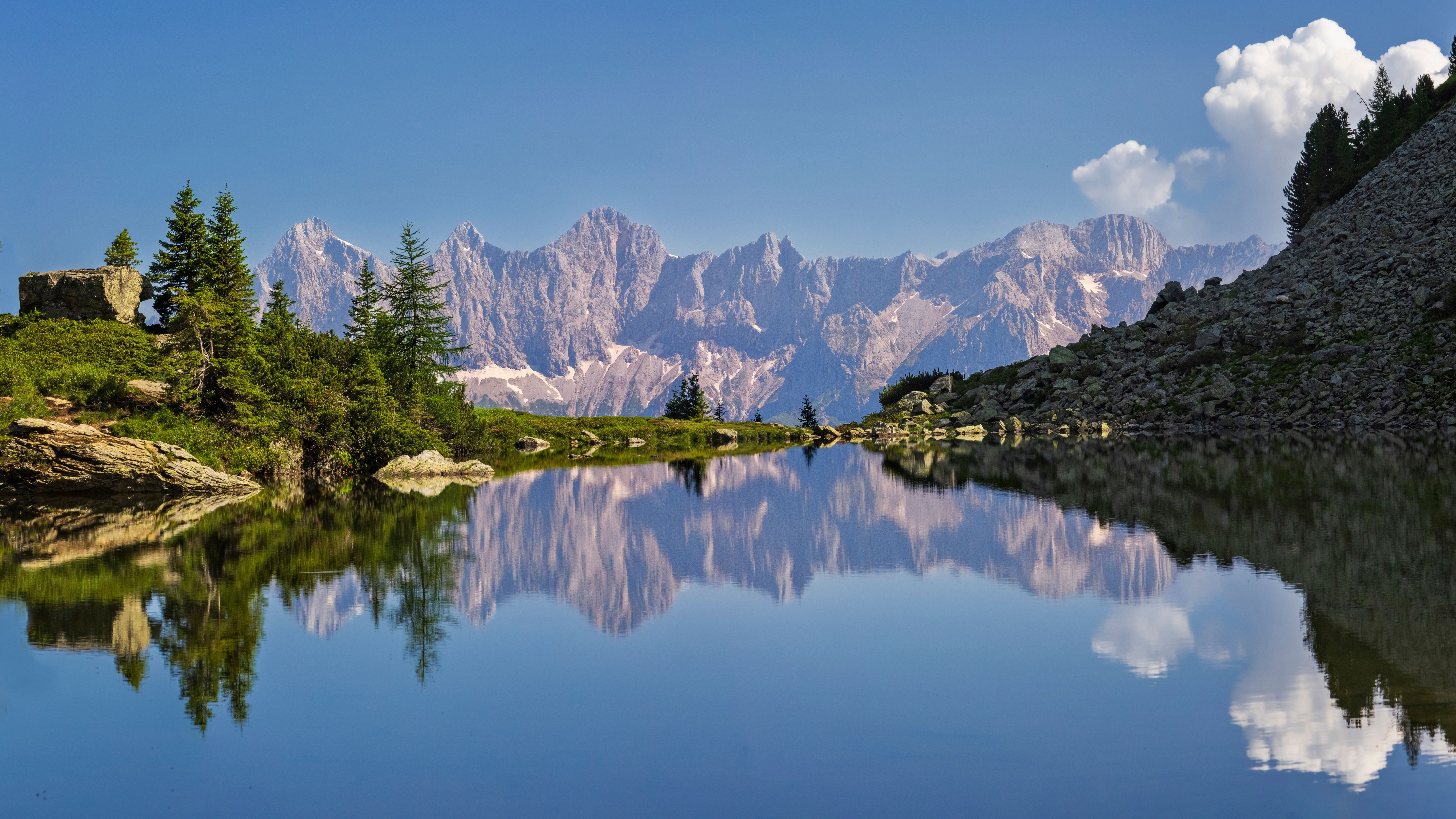 Klarer Bergsee mit Spiegelung der Dachsteinberge und Wolken, umgeben von Felsen und Bäumen.