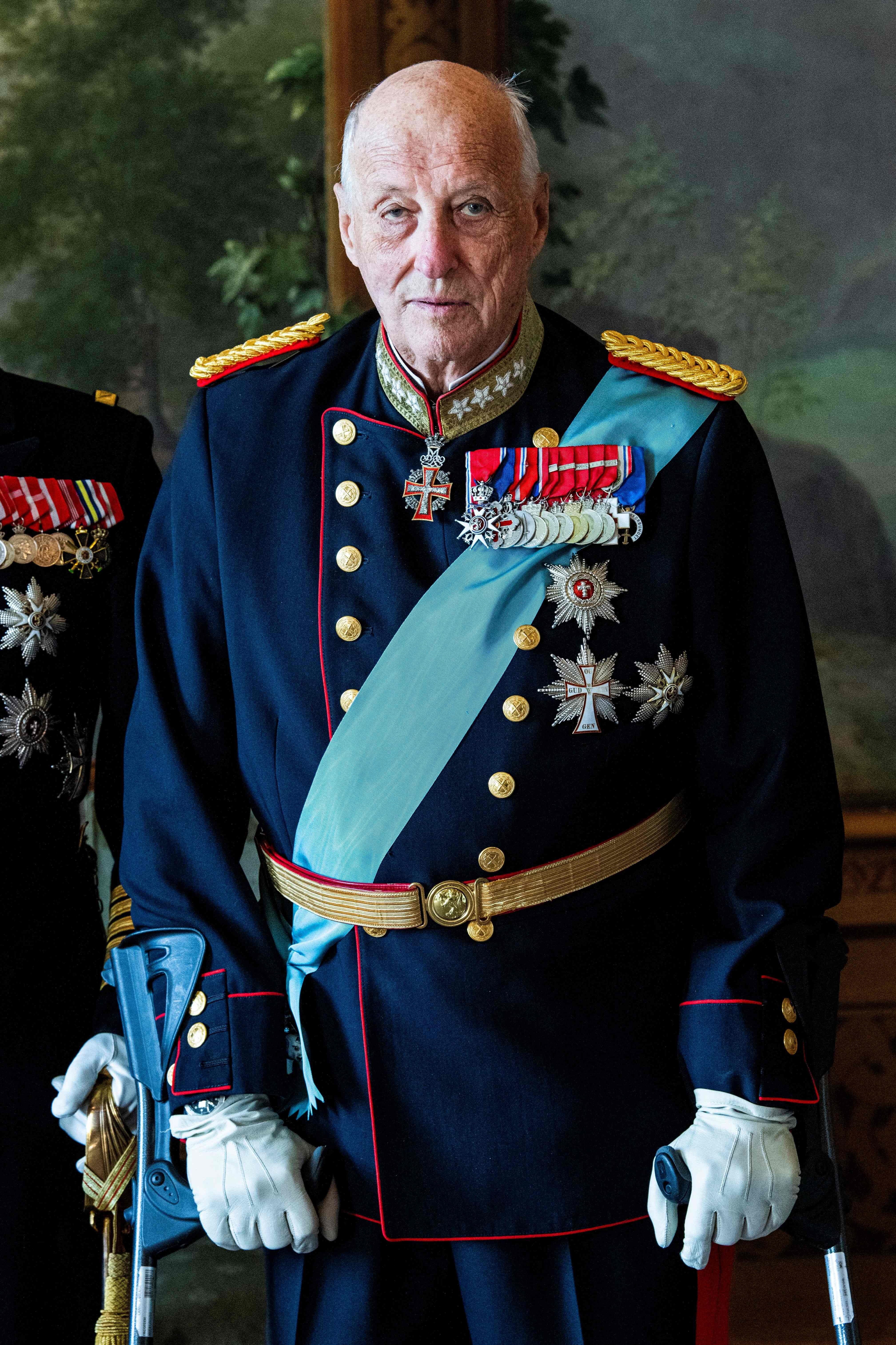 König Harald V. von Norwegen in Uniform mit Orden und Schärpe, stehend mit Gehhilfe.