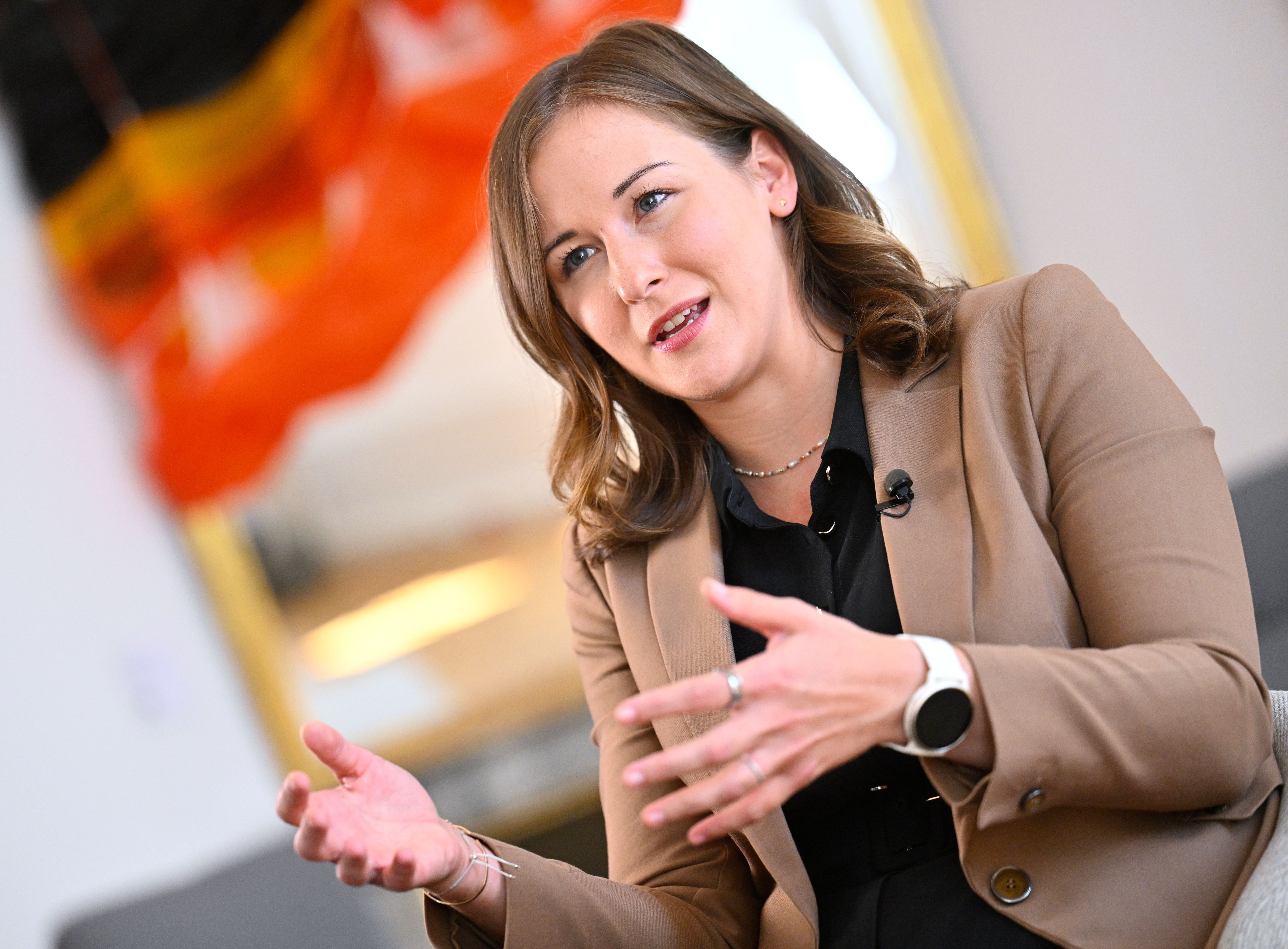 Frau in braunem Blazer spricht gestikulierend in einem Interview, unscharfe Flagge im Hintergrund.