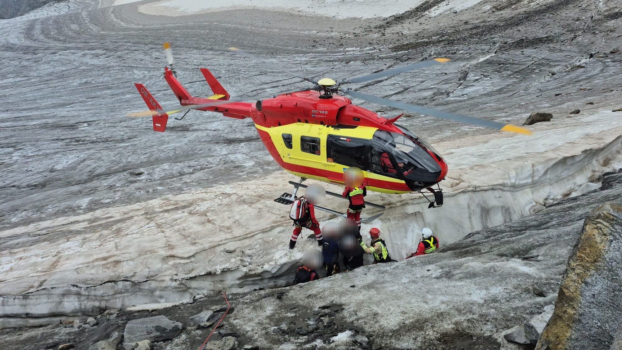 Ein Rettungshubschrauber fliegt nahe über einen Gletscher, Bergretter helfen Menschen aus einer Spalte.