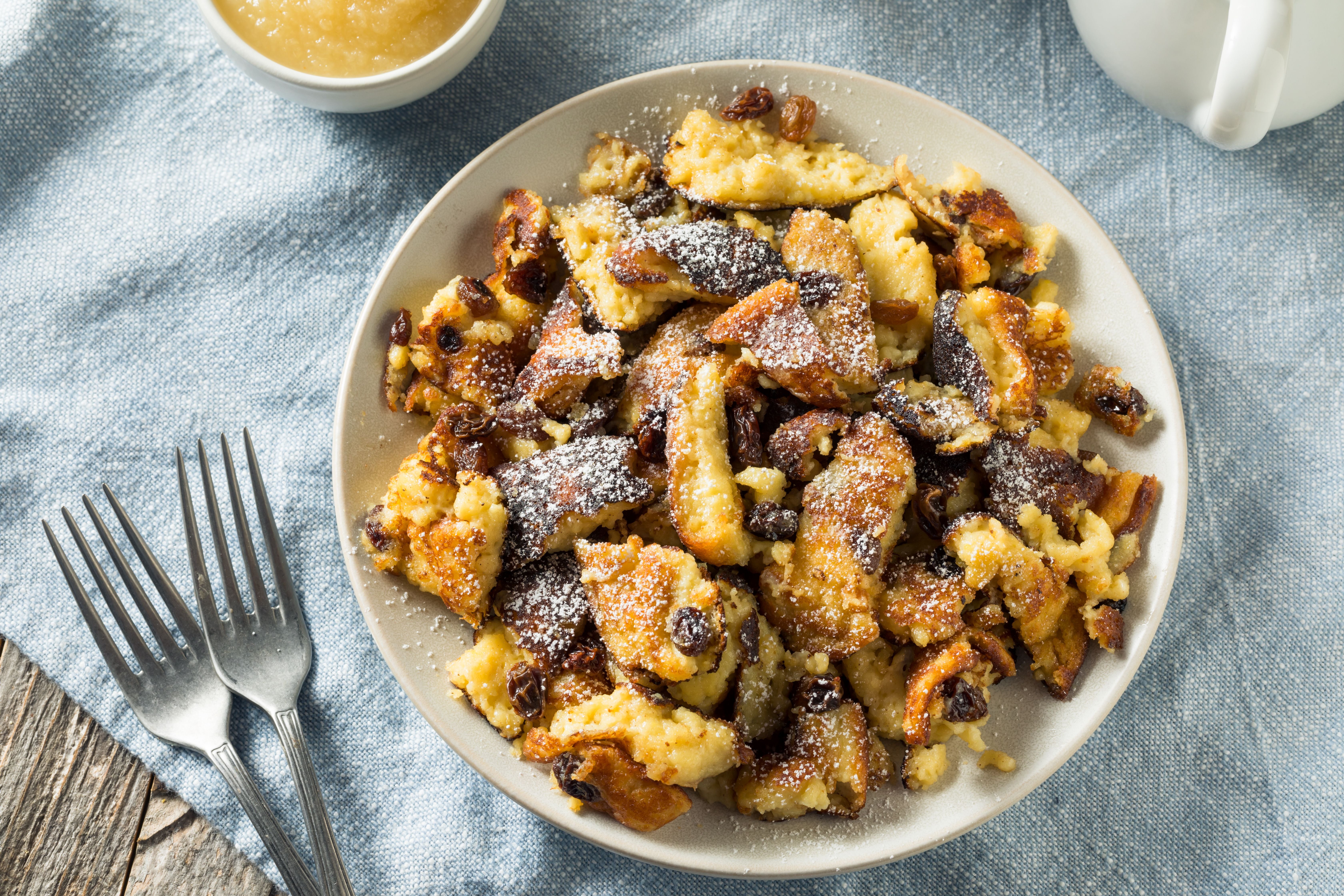 Kaiserschmarrn mit Rosinen und Puderzucker auf einem Teller, daneben Apfelmus und Gabeln.