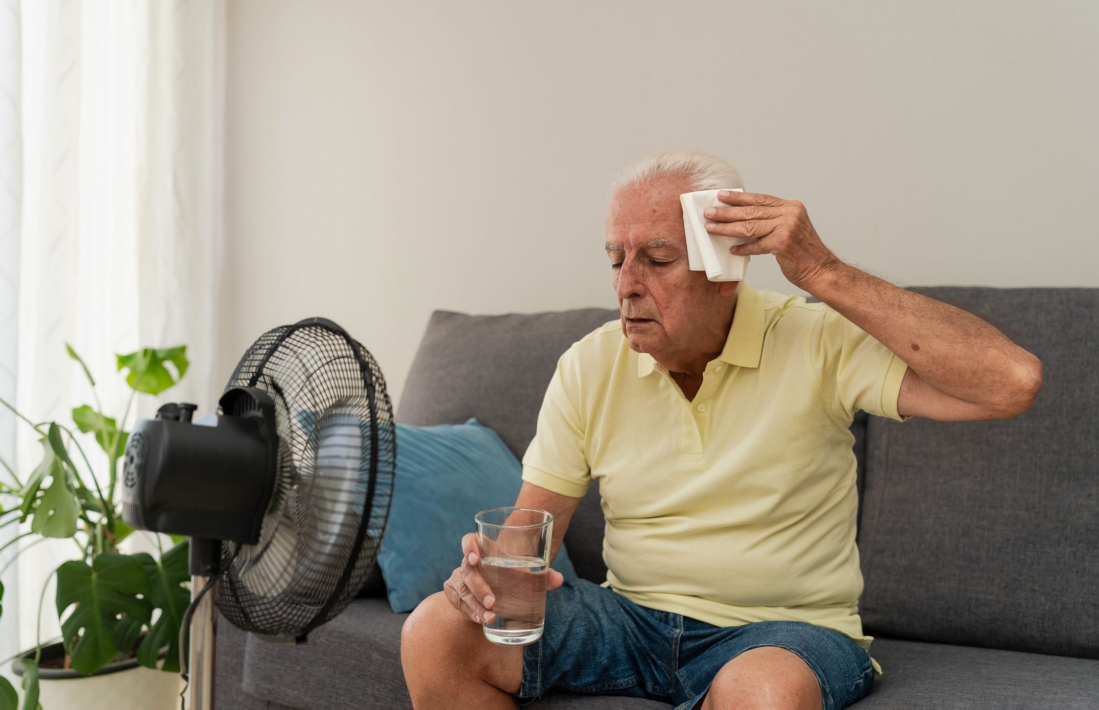 Älterer Mann sitzt auf dem Sofa, wischt sich Schweiß ab und hält ein Glas Wasser neben einem Ventilator.