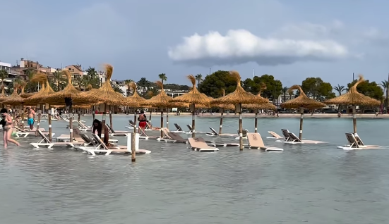 Sonnenliegen und Strohdächer stehen im überfluteten Strandbereich im flachen Wasser.