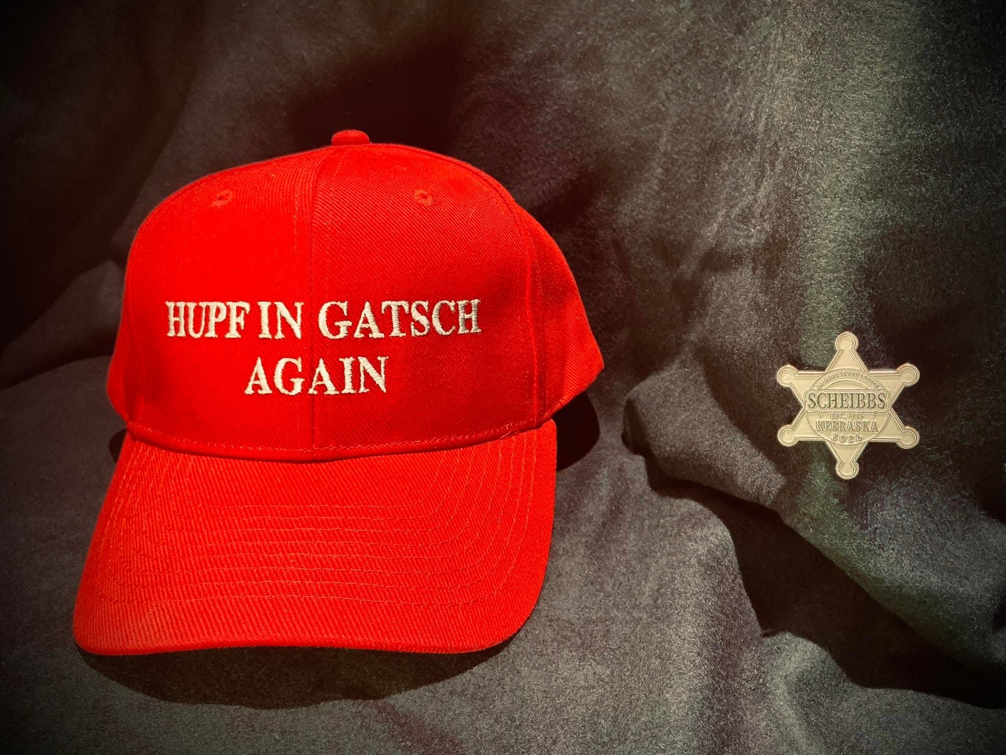Rote Kappe mit weißem Text „Hupf in Gatsch again“ neben Sheriffstern auf schwarzem Stoff.
