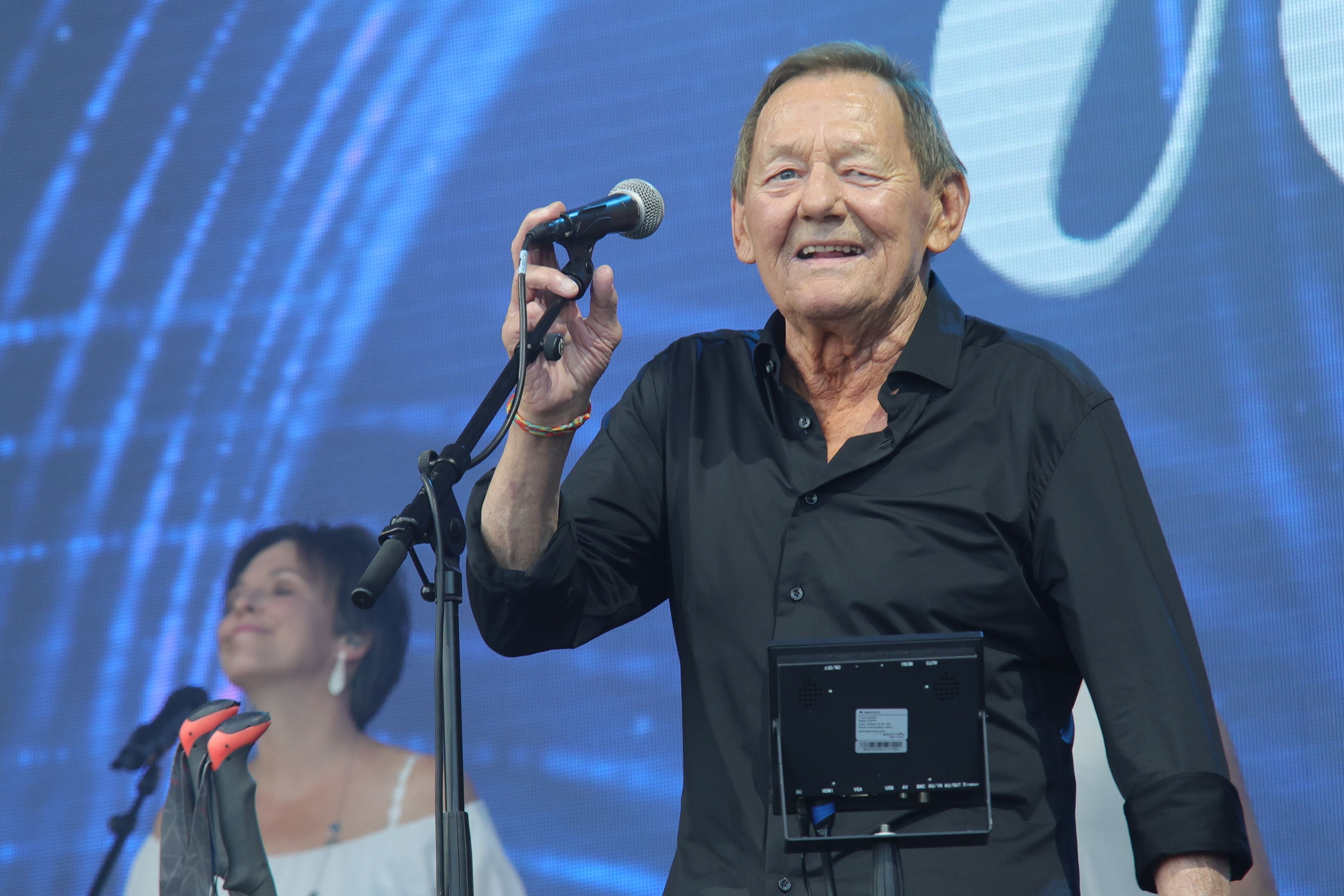 Ein älterer Mann singt auf einer Bühne ins Mikrofon, im Hintergrund eine Frau und blaue Beleuchtung.