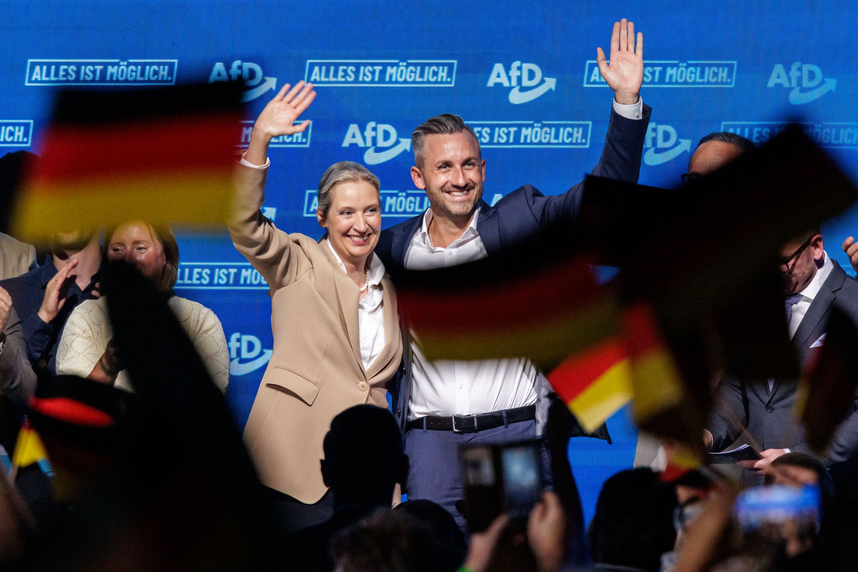 Zwei Menschen winken auf einer AfD-Wahlkampfveranstaltung, im Vordergrund deutsche Fahnen.