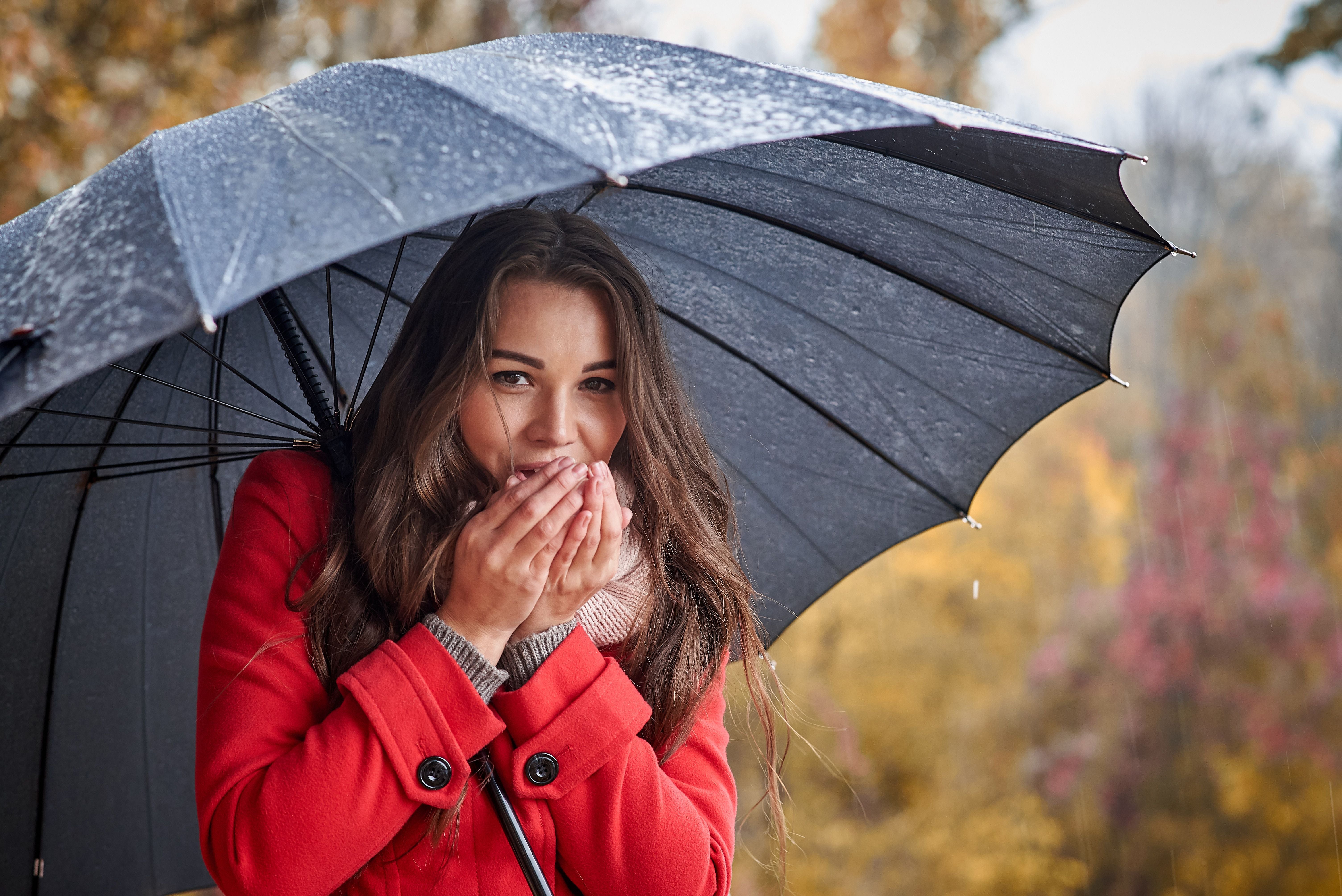 Frau im roten Mantel wärmt ihre Hände mit dem Atem unter einem Regenschirm im Herbst.